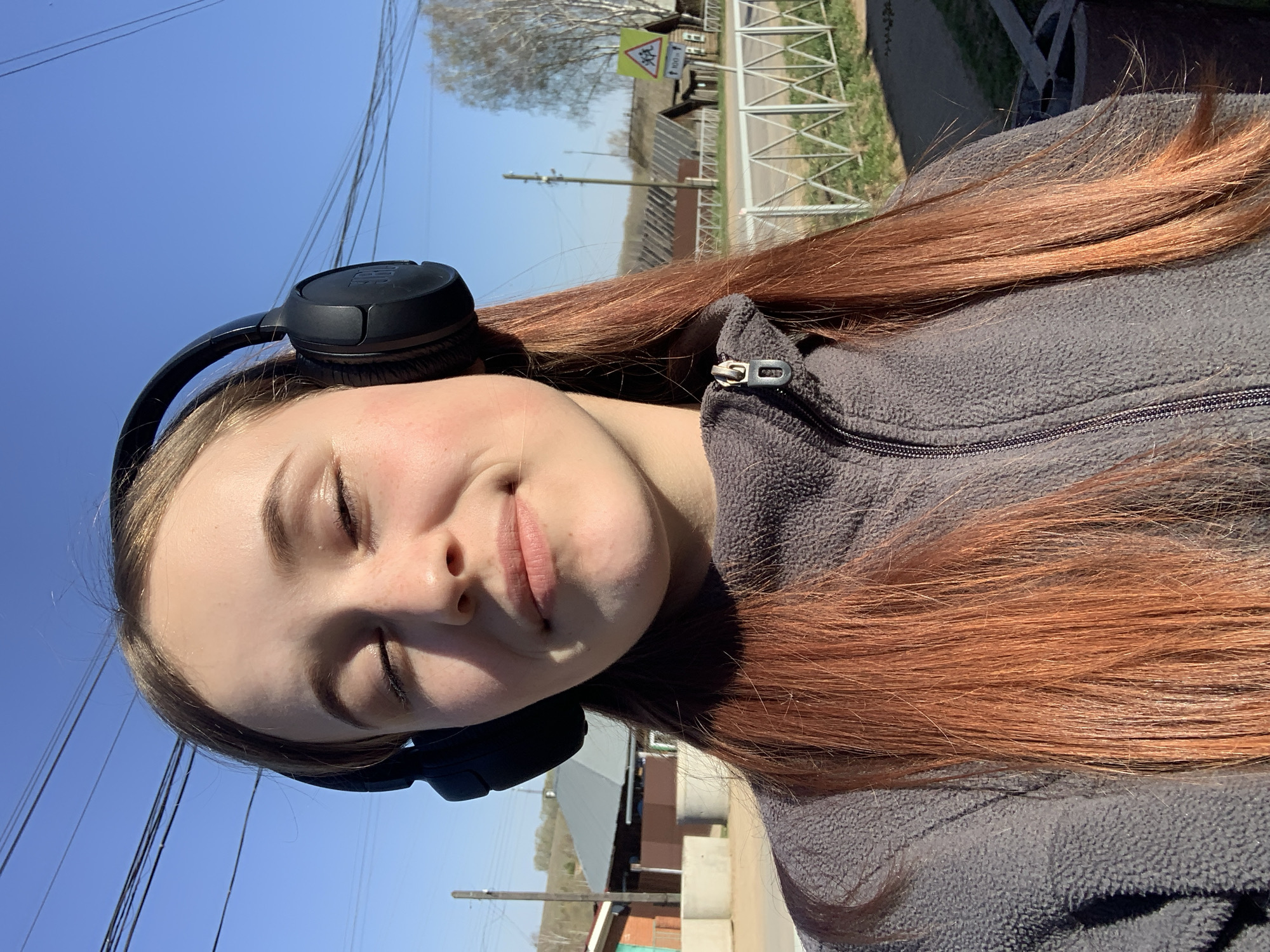 A young woman with auburn hair is facing the camera with her eyes closed. She is wearing black headphones and a gray zip-up jacket. The background features a blue sky, power lines, a road with a metal guardrail, buildings, and trees. A road sign indicating "Children" in Russian is visible. The scene appears to be outdoors during the daytime. The location is Фоки, Russia.