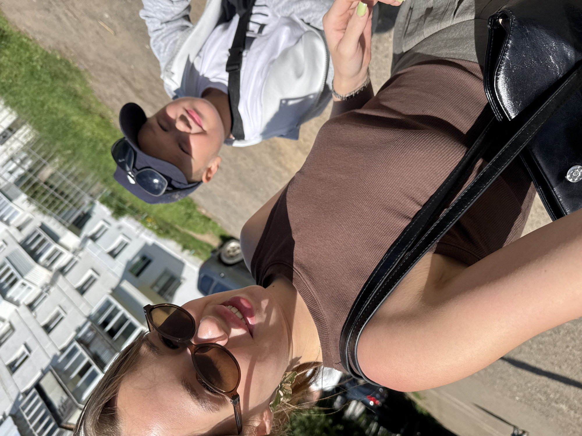 A selfie is captured outdoors on a sunny day in Chaikovsky, Russia, with a residential building in the background. A young woman in the foreground is wearing sunglasses and a brown tank top, with a black purse slung over her shoulder. Behind her, a young boy wearing a baseball cap and sunglasses is smiling. The environment appears to be a sidewalk or paved area adjacent to a grassy area and a building.