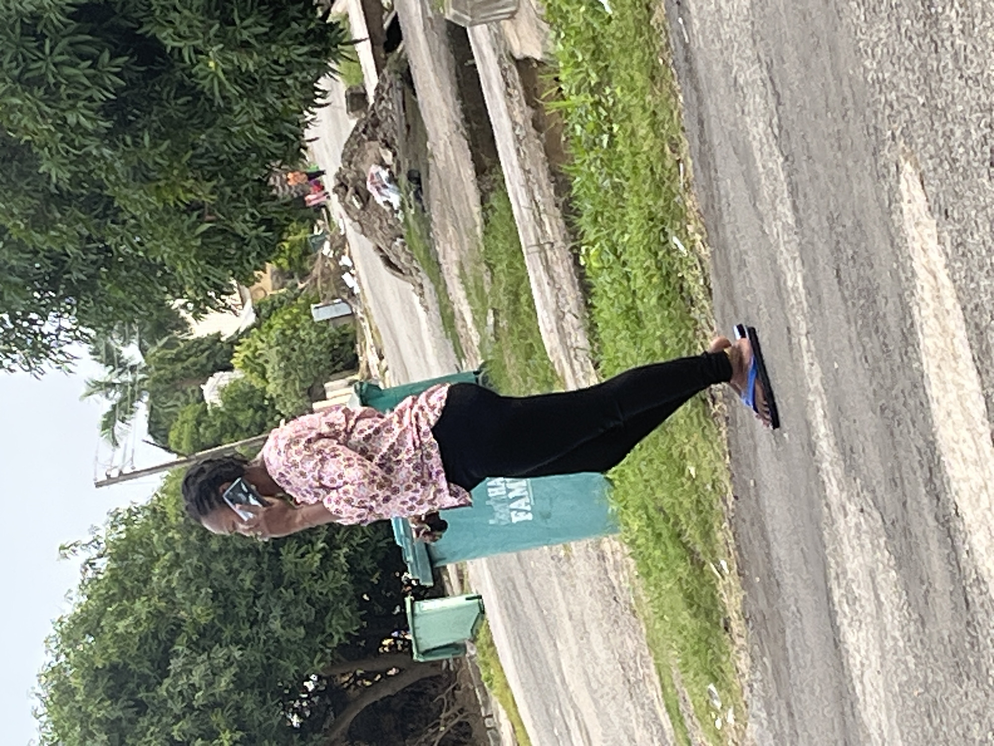 A woman is walking and talking on her phone on a paved road in Abuja, Nigeria. She is wearing a pink floral shirt, black leggings, and blue flip-flops. She is walking on the right side of the road, facing left. In the background, there are trees, two green trash cans, and a pile of debris. The sky is overcast.