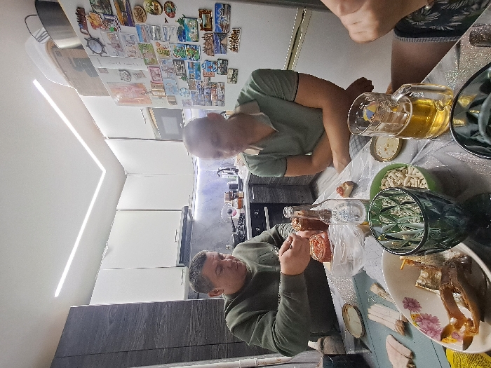 Three men are seated around a table laden with food and beverages. A glass of beer sits beside a green bowl containing peeled nuts. There are plates of various dishes, including fish. One man wears a green shirt, another a gray sweater. A fridge is visible behind them, adorned with decorative magnets. The room is lit by ceiling lights.