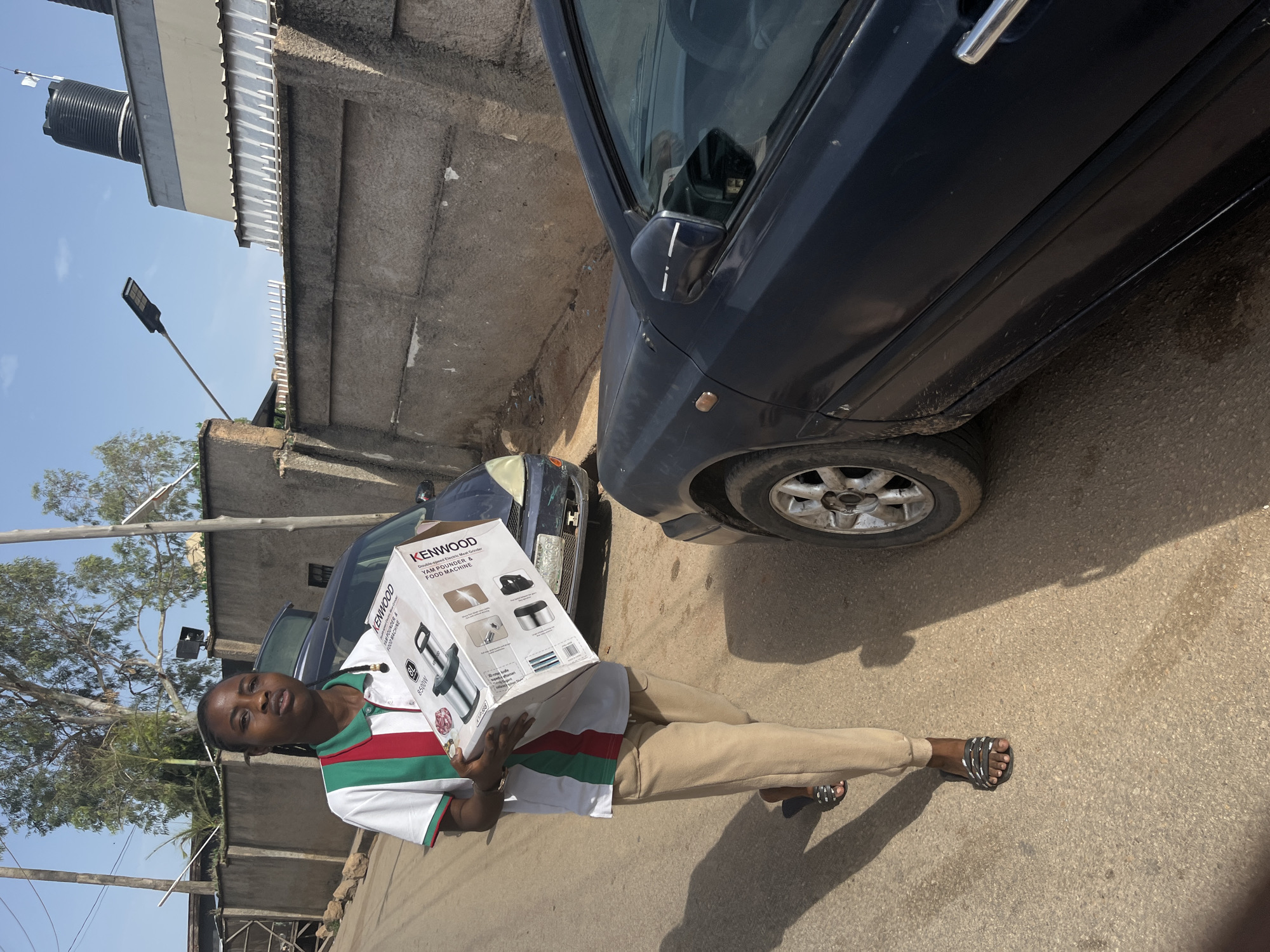 A young woman of African descent is walking on an asphalt road in Jos, Nigeria, carrying a Kenwood yam pounder and food machine box. She is wearing a white polo shirt with red and green stripes on the collar and sleeves, tan sweatpants, and black and white slide sandals. The woman is looking directly at the camera with a neutral expression. Behind her, a dark blue sedan is parked on the right side of the road, and another blue car is parked further down the street on the left. A concrete wall runs along the side of the road, with a street lamp and water tanks visible on top of a building in the background. The sky is clear and blue. The scene appears to be in a residential or commercial area with a mix of buildings and vehicles.