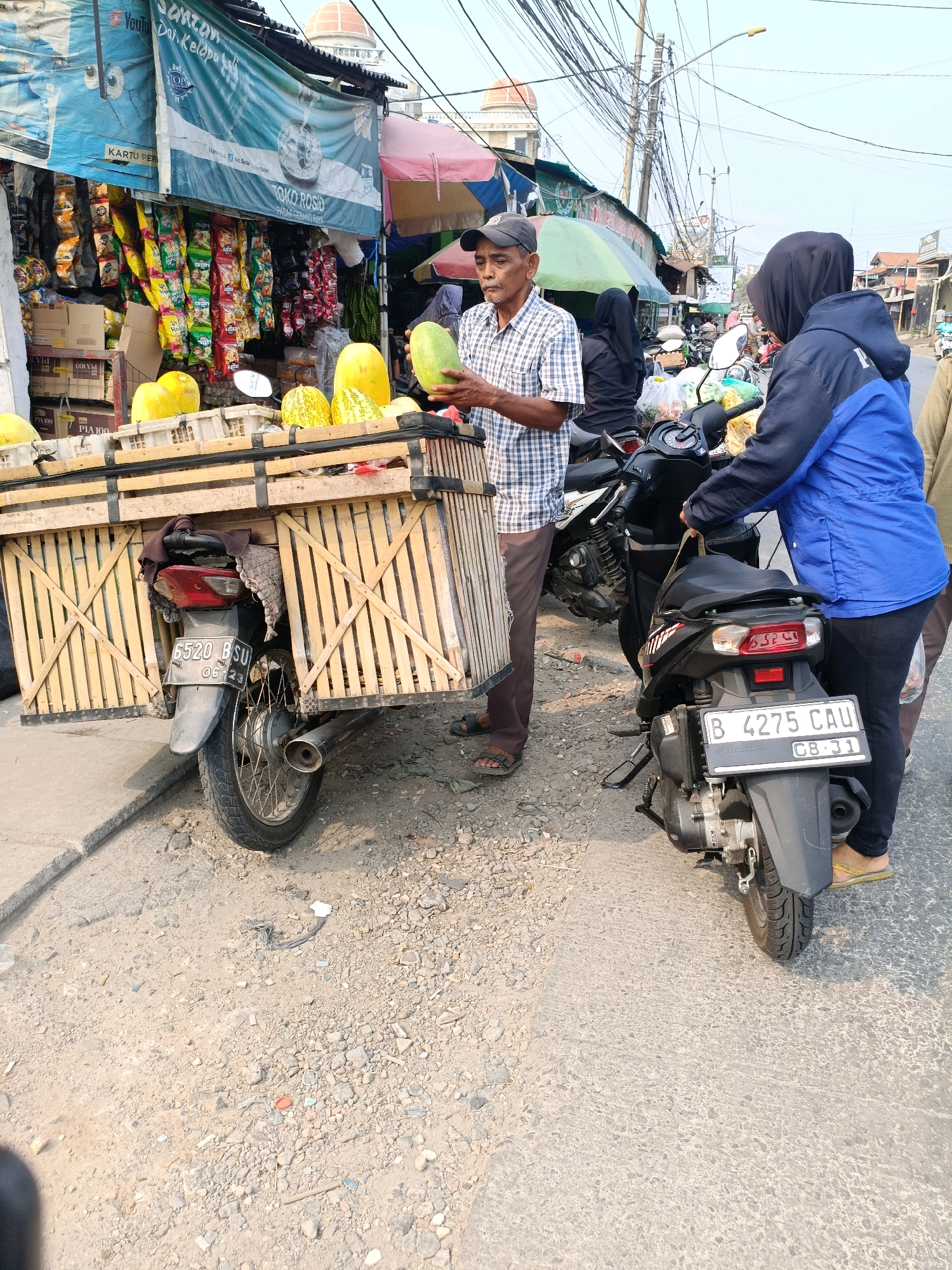 The image shows a street scene in Tangerang, Indonesia, likely a marketplace. In the foreground, a man stands next to a wooden cart filled with melons, which is attached to the back of a motorcycle. He is holding a melon in his hands. Behind him, a woman wearing a blue jacket and a black hijab is standing next to another motorcycle. A storefront with banners and merchandise is visible in the background. The environment appears to be a busy street with power lines and buildings.