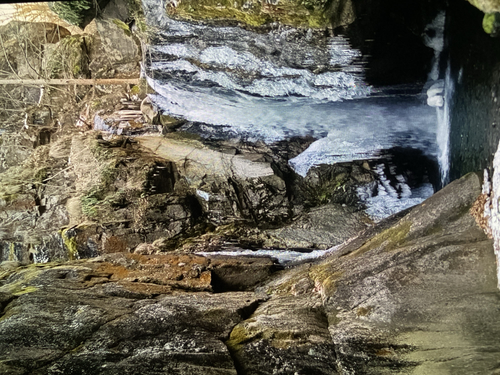 The image displays a partially frozen waterfall or stream cascading through a rugged, rocky gorge. The scene is dominated by dark, textured rock formations, many of which are covered in patches of green moss. Bare tree branches, characteristic of winter, extend from the upper sections of the gorge, adding to the desolate yet natural ambiance.
The water flows down in several stages, with significant ice formations clinging to the rocks and partially obscuring the movement of the water. These ice structures appear as undulating, frozen cascades, reflecting a cold weather event, likely during winter or early spring. At the base of the main waterfall, the water collects in a dark, shadowy pool, with additional ice visible along its edges, suggesting a deep, secluded area.
No people or animals are visible in the photograph. The lighting is diffused, suggesting an overcast day or the deep shadows of the gorge itself, emphasizing the cool tones of the ice and rocks. The overall impression is one of natural ruggedness and cold beauty. There is no visible text in the image. The location is specified as Bankya, Bulgaria.