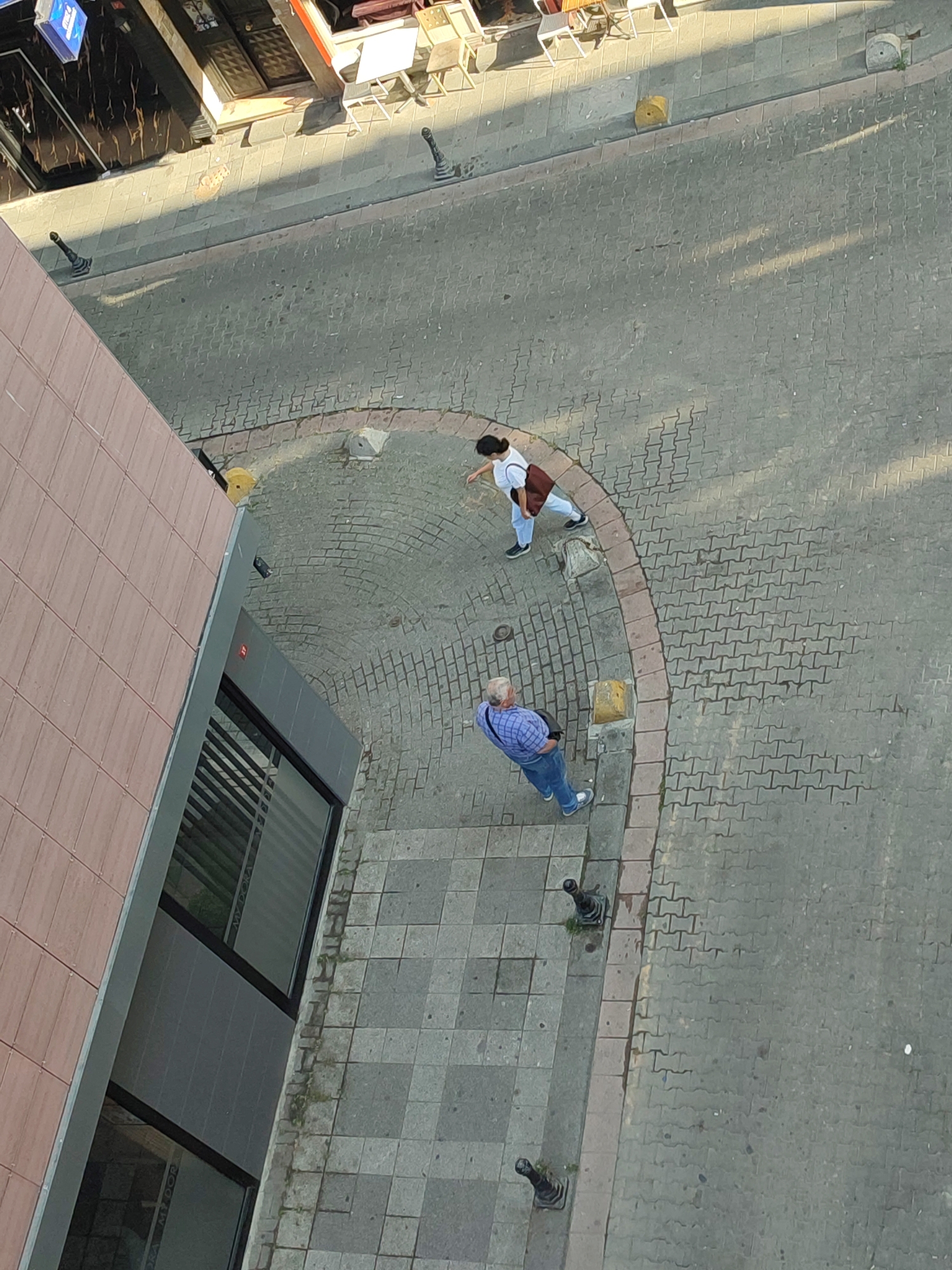 The image shows an aerial view of a street scene in Kadıköy, Turkey. Two individuals are visible on a cobblestone sidewalk adjacent to a building. In the foreground, a man with gray hair wearing a blue checkered shirt, jeans, and sneakers is standing and looking down. Behind him, a woman with dark hair, wearing a white top, light-colored pants, and a brown shoulder bag, is walking away from the camera. The street is paved with interlocking bricks, and there are some decorative bollards along the curb. Sunlight casts shadows across the scene, indicating it is daytime. The building to the left has a textured facade and large windows. The overall environment suggests a commercial or residential urban area.