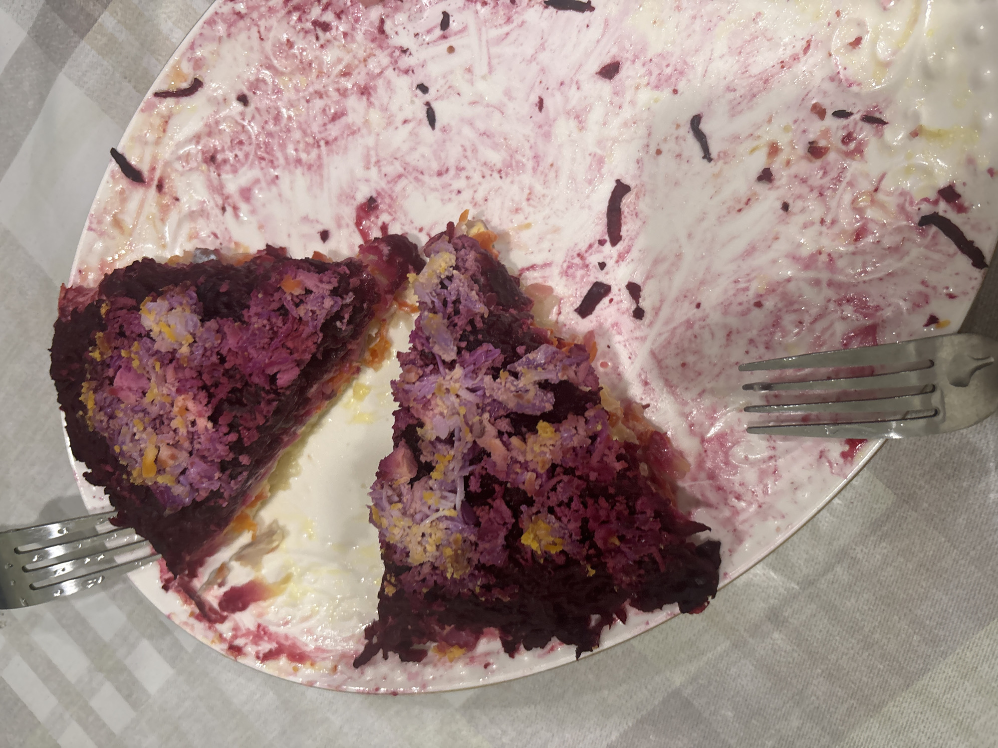 The image depicts two slices of a layered salad, likely "Herring Under a Fur Coat" (Селёдка под шубой), on a white plate with pink and red stains from the beetroot. The salad consists of layers of dark red beetroot, lighter colored ingredients like mayonnaise and perhaps carrots or potatoes, and a purple, shredded topping which may be beetroot or cabbage mixed with dill and possibly egg or cheese. Small pieces of grated yellow and purple material are visible on top of the salad. A fork is present on the left side of the plate, and another fork is partially visible at the bottom. The plate is set on a light grey and white checkered tablecloth. The background is out of focus. The scene is located on a table in a dining setting. There is no information to suggest the image is from Sinyavino, Russia.