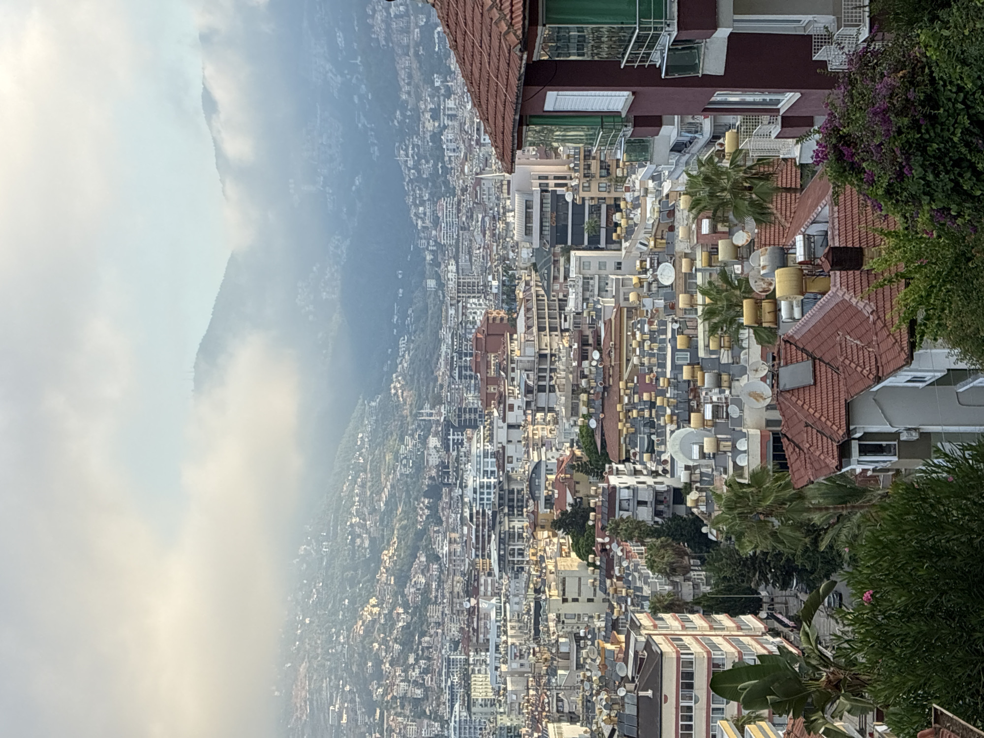 The image shows a panoramic view of Alanya, Turkey, captured from a high vantage point. The cityscape is dense with buildings, many featuring red tile roofs and solar water heaters on their rooftops, characteristic of the region. The buildings vary in height and architectural style, with some exhibiting modern designs and others appearing more traditional. In the background, lush green hills rise, partially obscured by a soft layer of clouds that also cast a gentle haze over the scene, suggesting either early morning or late afternoon lighting. Trees and other vegetation are interspersed throughout the urban landscape, and a few palm trees are visible, adding to the Mediterranean feel. The sky is a pale blue, with the clouds creating a soft, diffused light. No people or specific actions are discernible in the image. The overall impression is one of a bustling coastal city nestled against a natural landscape.