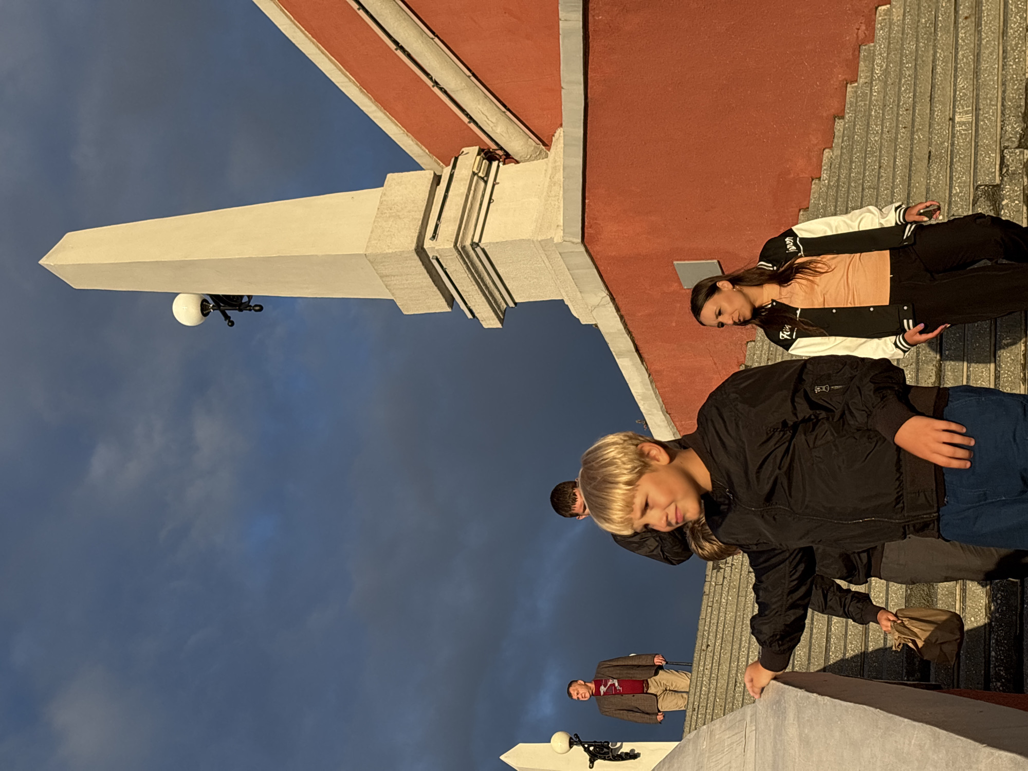The image shows a group of people on a wide staircase leading up to a red-bricked building with a white obelisk structure. The sky is dark with clouds. A young boy with blonde hair in a black jacket is in the foreground, looking to his right. Behind him, a woman in a black and white jacket and an orange shirt is walking down the stairs. Further up the stairs, a man in a brown jacket and a red shirt is standing and looking to his left. Another man in a dark jacket is visible behind the blonde boy. The staircase appears to be made of concrete or stone. The setting is outdoors, likely in a public space in Nizhny Novgorod, Russia, given the architectural style which is reminiscent of monuments in that city. The lighting suggests it might be late afternoon or early evening, with sunlight illuminating parts of the scene.