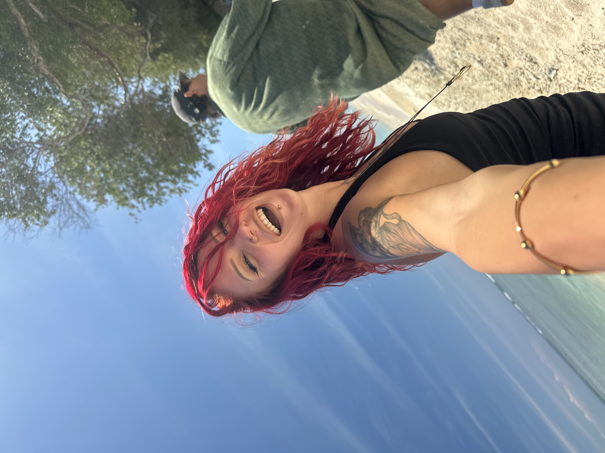 The image shows a woman with bright red hair, wearing a black tank top, and a gold bracelet on her arm. She is smiling and has a tattoo on her upper arm. She is standing on a beach with blue sky above. In the background, a person wearing a green robe and a dark helmet stands under a tree. The location is Unknown, Indonesia.
