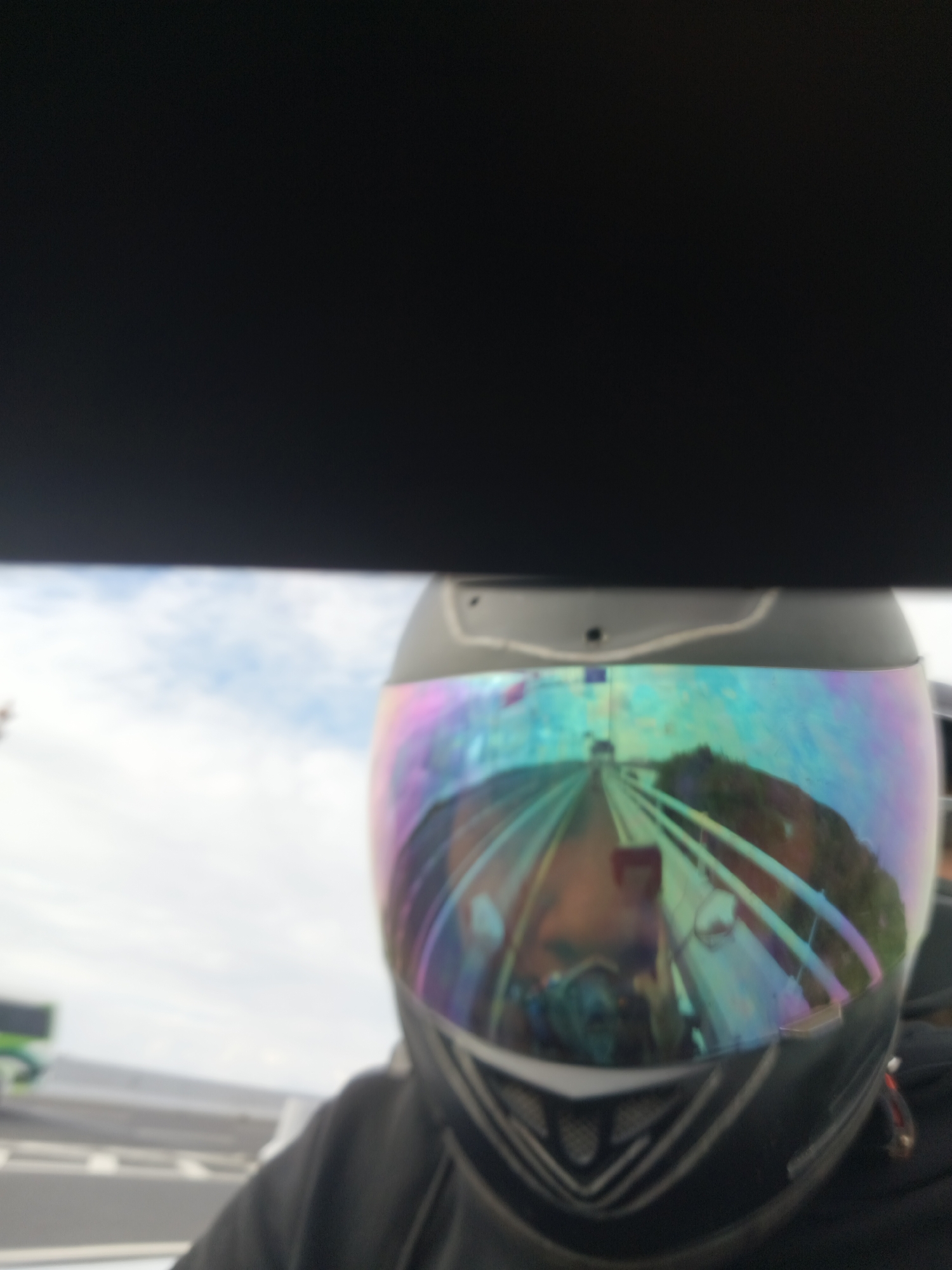 The image shows a first-person perspective of someone wearing a black motorcycle helmet with a reflective, iridescent visor. The visor displays a distorted reflection of a road ahead, possibly a highway or a bridge, with some greenery on the sides. A portion of a person's face is visible in the reflection. In the background, the sky appears overcast with clouds. Part of a black structure, likely a car's dashboard or roof, is visible at the top of the image, obscuring the upper portion of the scene. There is no clear indication of the city of Kuta, Indonesia, within the image content.
