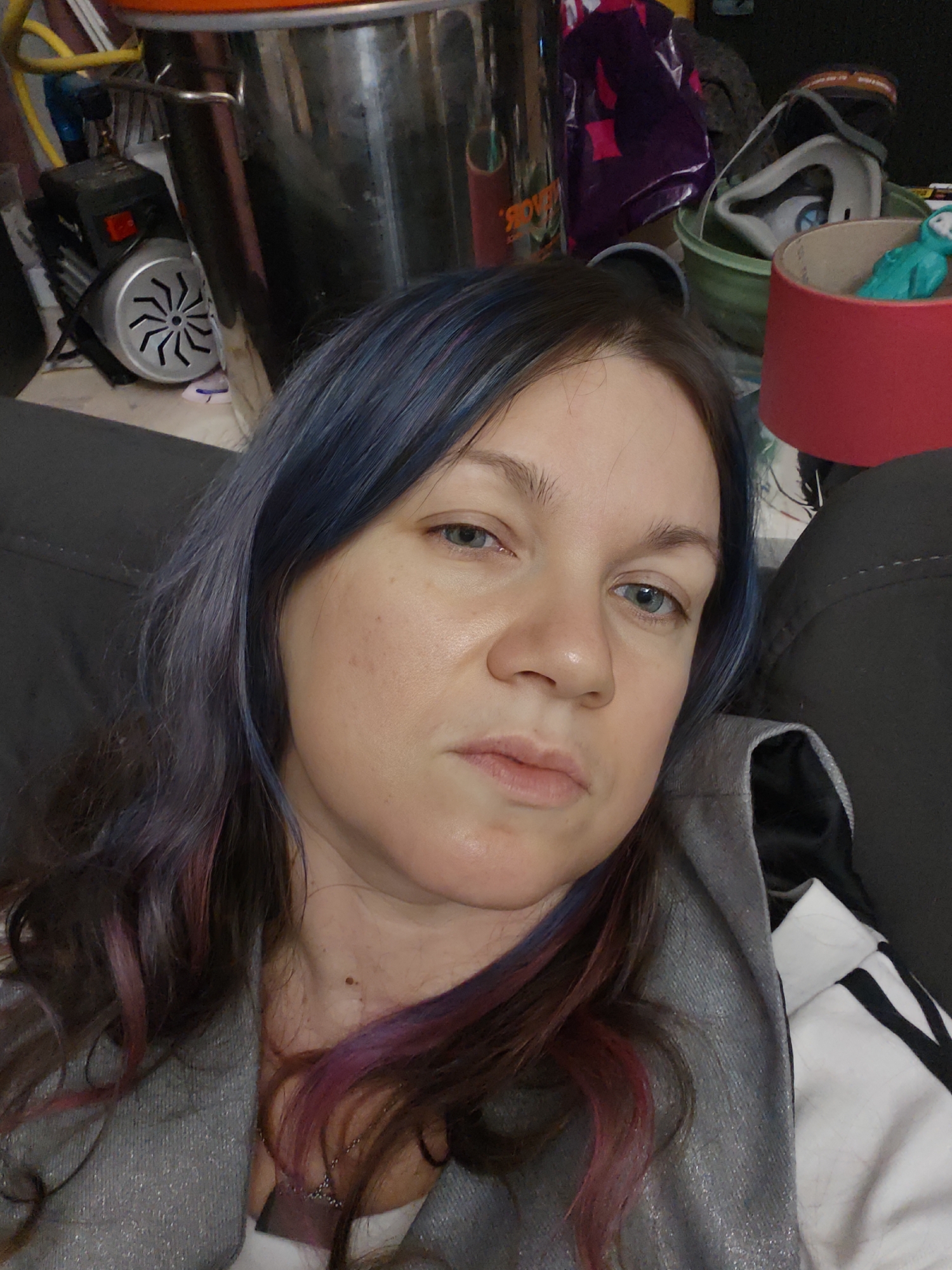 A selfie taken indoors shows a woman with blue and pink-streaked hair. She has fair skin and light-colored eyes. She is wearing a gray hooded sweatshirt with a white and black sleeve visible. The background is cluttered. Visible items include a large metal pot, a black motor with a fan, a red lampshade, and assorted containers and items obscured by shadows and poor lighting. The location is unspecified except that it is in the city of Свистуха (кузяевский С/о), Russia, which only specifies a region.