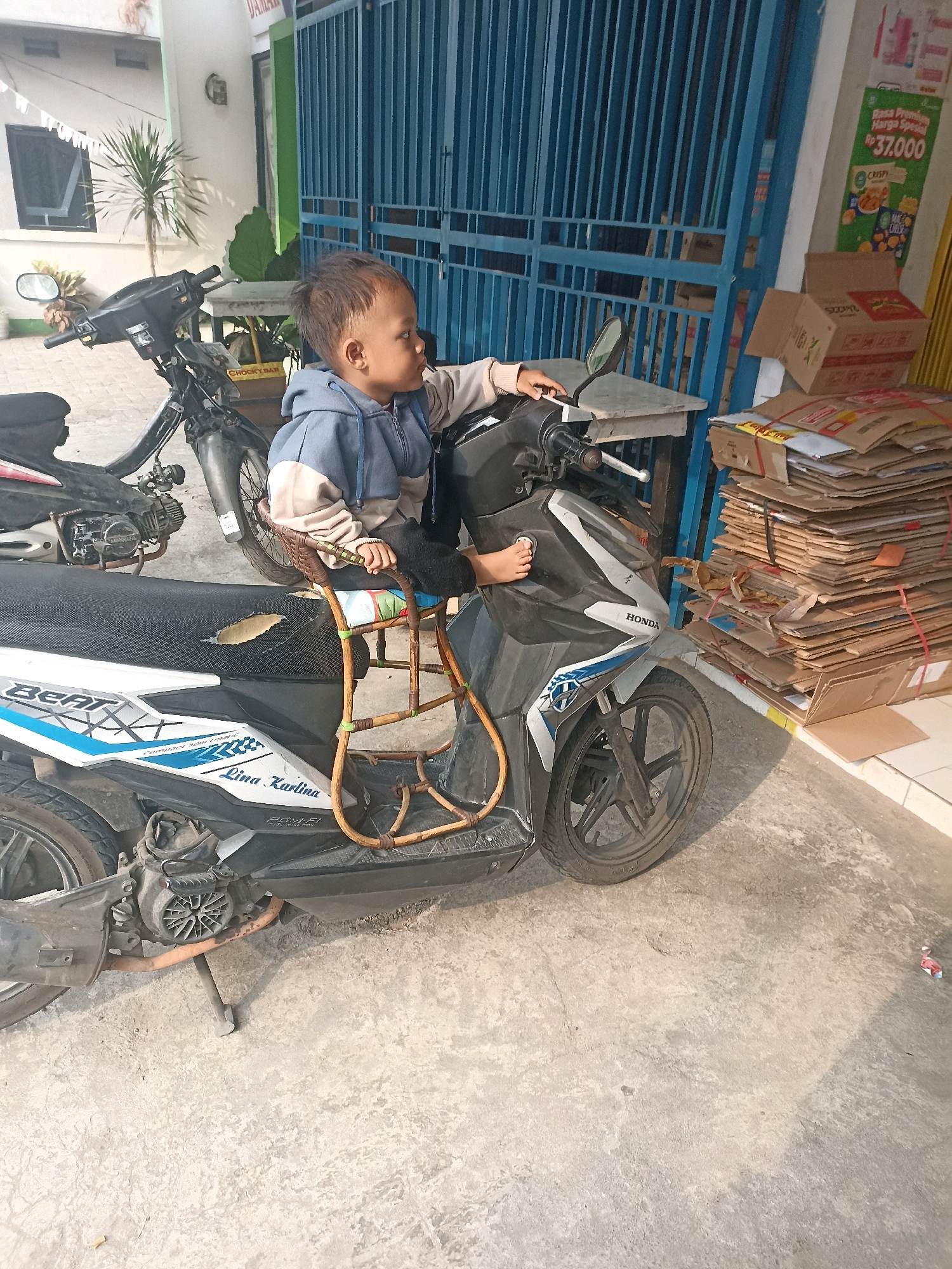 A young child is seated on a child seat attached to the back of a black and white Honda Beat scooter. The child is wearing a blue and beige hooded jacket and appears to be looking to the right. The scooter is parked on a concrete surface, possibly a sidewalk or driveway, in front of a blue metal gate. To the right of the scooter, a large stack of cardboard boxes is piled up, some of which are tied with red string. In the background, there is a white building with a window, a table, and some greenery. A second motorcycle is partially visible to the left of the main scooter. The scene suggests an outdoor setting in a residential or commercial area.