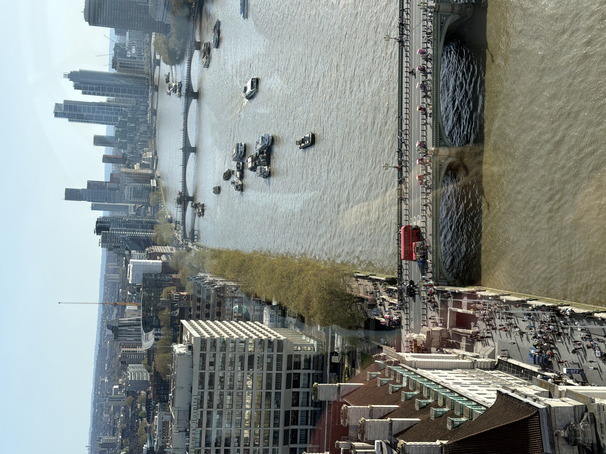 This is an elevated view of London, United Kingdom, featuring the River Thames. Several bridges span the river, including what appears to be Tower Bridge, recognizable by its distinct architectural design. The river is active with boats. Alongside the river, there is a pedestrian area bustling with people, suggesting a popular tourist or recreational area. Buildings of various heights and architectural styles dominate the cityscape. The image captures a mix of modern and older constructions. The weather appears clear, with good visibility. The presence of numerous people on foot indicates a lively urban environment.