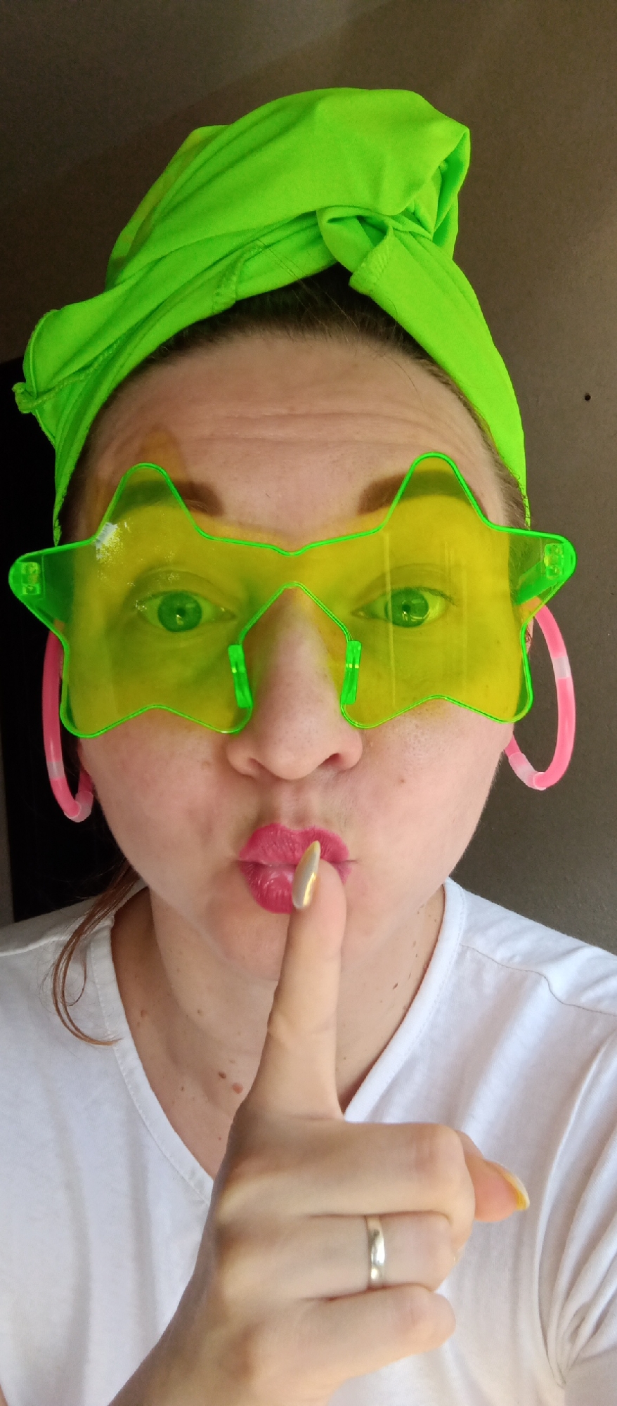 A person is shown from the chest up, holding a finger to their lips in a gesture to be quiet. The person has a bright green headband, large green star-shaped glasses, and pink hoop earrings. They are wearing a white t-shirt. A gold-colored manicure is visible on the finger being held to the lips. A simple silver ring is worn on another finger. The location is unknown.