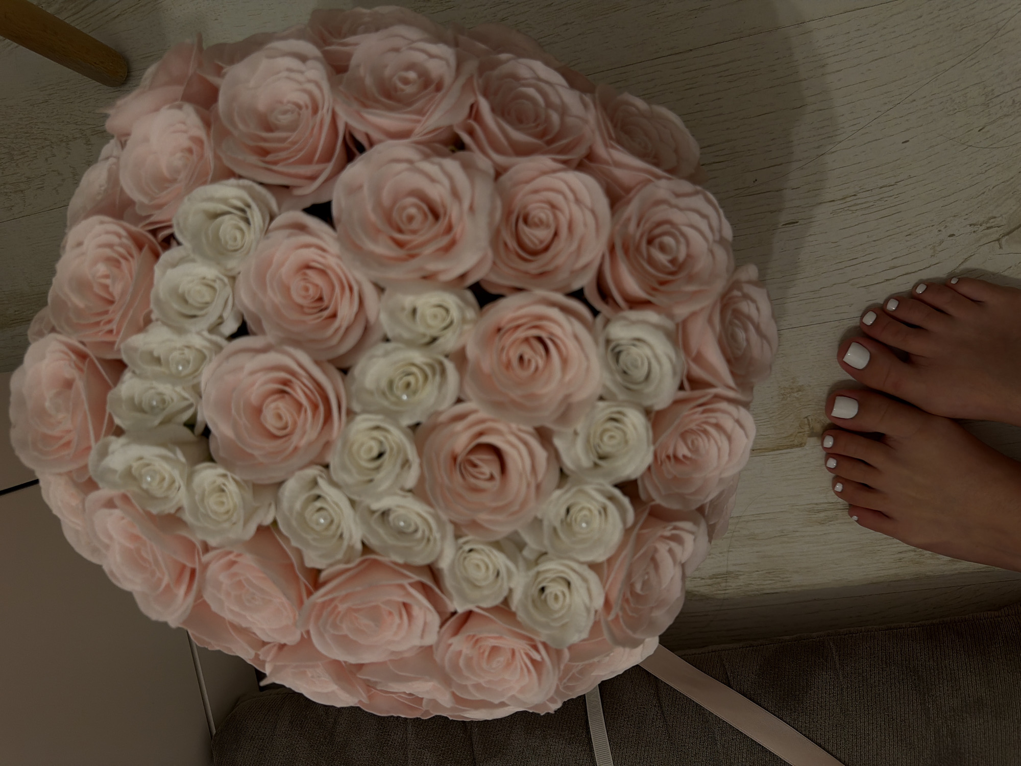 An overhead shot shows a large bouquet of light pink and white roses in the center-left of the frame, with a woman's feet to the right. Her toenails are painted white. The bouquet sits on a grey couch or cushion with a light pink ribbon. The background is a light-colored wooden floor. There are no clues to indicate this photograph was taken in Plovdiv, Bulgaria.