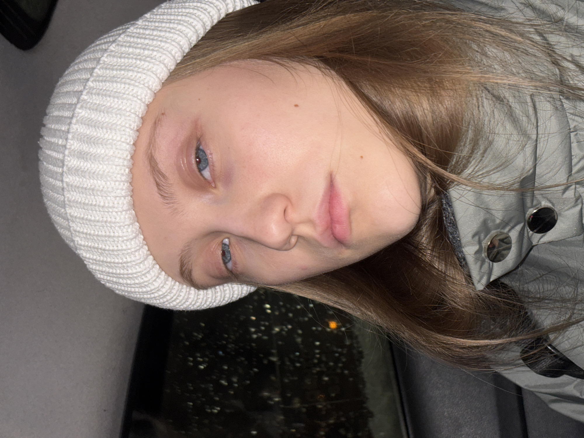 A young woman with light skin, blue eyes, and light brown hair is wearing a white knit beanie and a gray jacket with silver buttons. The image is taken indoors, likely inside a car, as blurred lights are visible through the window behind her. The location is potentially Perm, Russia.