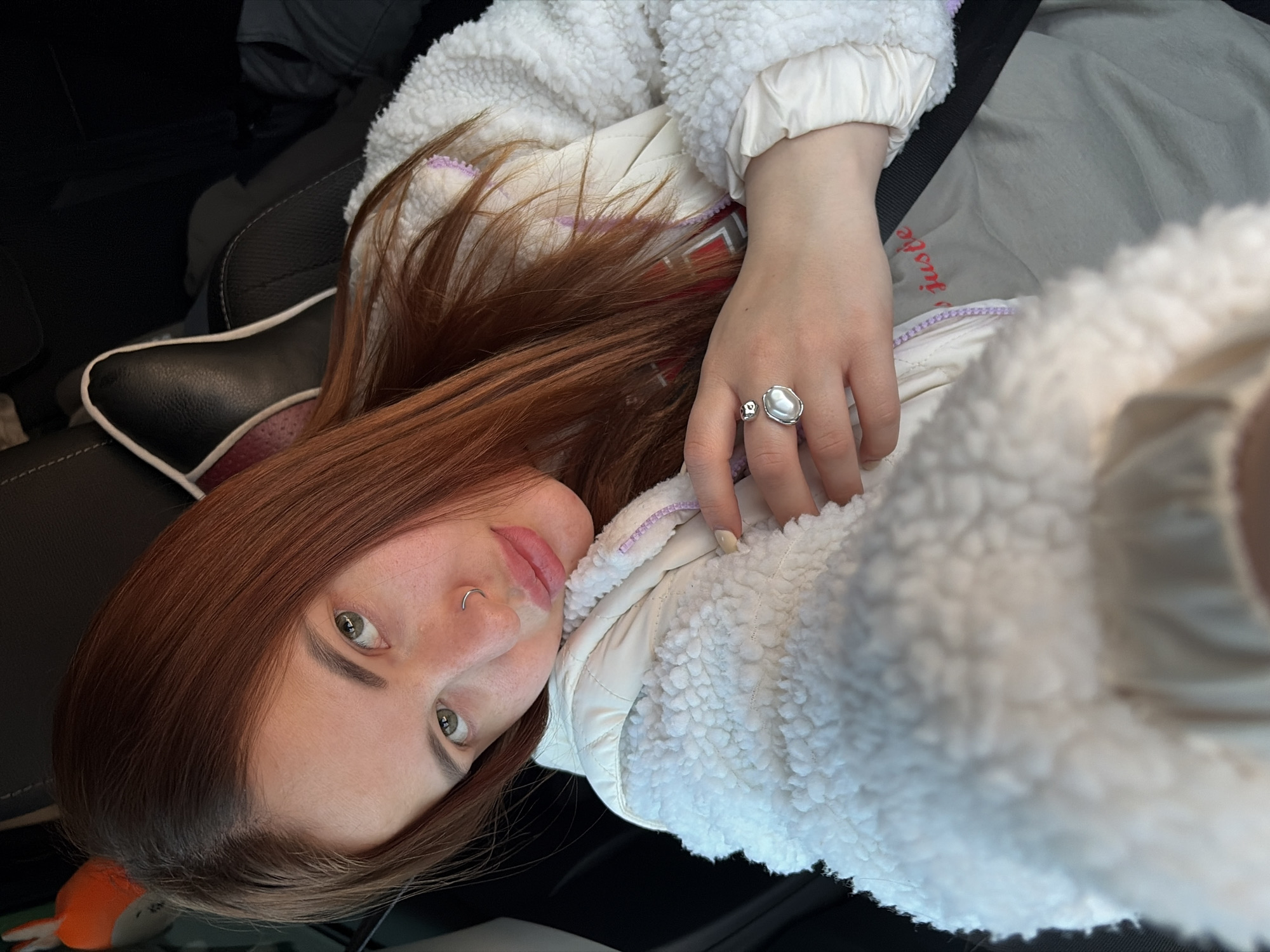 A young woman is depicted in a close-up, upward-angled selfie, positioned inside what appears to be a car. She has long, reddish-brown hair and green eyes, with a septum piercing (nose ring). Her lips are slightly parted in a neutral expression. She is wearing a white, fluffy, sherpa-style jacket with a light-colored, possibly lilac, zipper lining, over what seems to be a lighter-toned underlayer. A prominent silver ring with an irregular, pearl-like or organic-shaped white stone is visible on her left ring finger. Her fingernails are painted a light, neutral color.
The setting is an interior, characterized by dark car seats with white piping and a visible black seatbelt draped across her body. The background is dark and blurred, suggesting the confined space of a vehicle. A light-colored fabric with faint red text, possibly reading "juste," is partially visible behind her arm. An orange, plush object can be glimpsed in the lower left background, near what looks like a headrest. The lighting suggests daytime, but the source is diffuse, possibly from inside a car or an overcast sky. The overall activity is a self-portrait or a casual photo taken while seated in a car, likely in Alexandrovka, Russia.