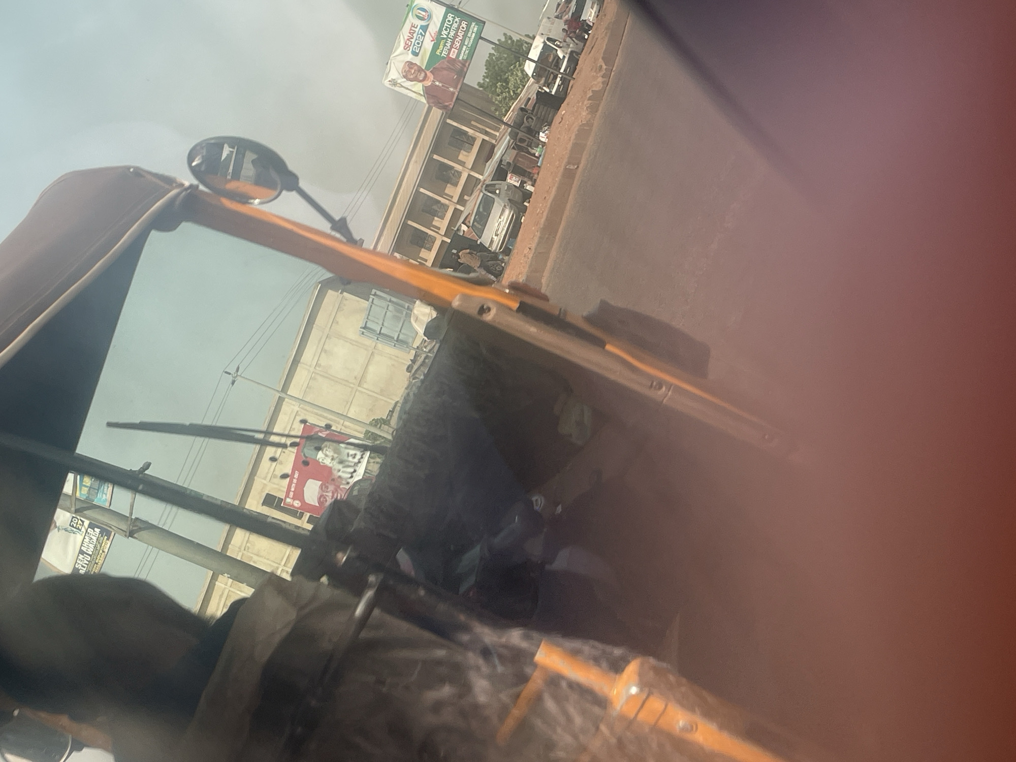 The image, taken from inside a vehicle, likely an auto-rickshaw, captures a street scene in Keffi, Nigeria, on what appears to be a clear, bright day, possibly late morning or afternoon. The perspective is tilted, suggesting movement or a quick shot, and a large reddish-orange flare obscures the right side of the frame.
In the foreground, parts of the vehicle's interior are visible: a dark brown soft top or canopy on the left, an orange frame element stretching across the middle, and a side mirror reflecting indistinct shapes. Windshield wipers are also seen across the top left portion of the view outside. A dark, blurry form of a person or object is visible in the bottom left, inside the vehicle.
The background reveals a bustling street with buildings, other vehicles, and pedestrians. A light-colored building with multiple windows stands prominently. A paved road or shoulder runs alongside, with parked and moving cars, including a white sedan. Utility poles and wires crisscross the sky.
A large political billboard dominates the upper middle, featuring a smiling man identified by the text as "Pharm. VICTOR TERAH PATRICK SENATOR" for "SENATE 2027." Smaller text indicates "KADUNA SENATORIAL DISTRICT, KADUNA STATE." Another, partially obscured billboard on the lower left displays "SEN AHMED ALIYU WADADA" and "WITH US-2027," along with "EFU WAMEBA." The OCR also detected "29" and "600," possibly part of this or another adjacent sign. The visible text strongly points to an election campaign in Nigeria.