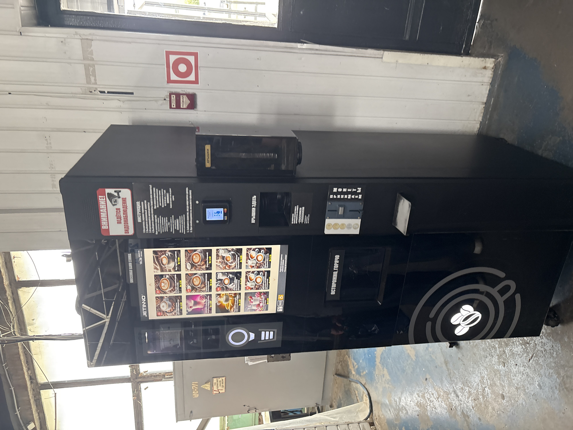 The image shows a black, freestanding coffee vending machine, likely located indoors in an industrial or commercial setting. The machine has a large display screen showing various coffee options with prices. Below the screen, there are coin slots and a dispensing area. To the left of the machine, there is a grey electrical panel with Cyrillic text and a warning sign. The background appears to be a concrete wall with metal and glass elements, possibly indicating a workshop or a public building. The floor is concrete. A fire alarm is visible on the wall to the right of the vending machine. The overall impression is a functional, utilitarian space.