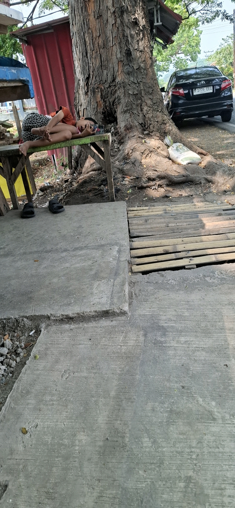 A person is lying on a green-painted wooden bench under a large tree in Cisalak, Indonesia. The person, who appears to be a child, is wearing a short-sleeved orange shirt and patterned shorts. They are lying on their back, with their head turned to the side and resting on their hands, looking at a smartphone in their hands. The bench is positioned on a concrete surface, with a wooden walkway made of planks to the right of the bench. A black sedan is parked on the road behind the tree. The environment appears to be outdoors, with natural vegetation visible in the background.