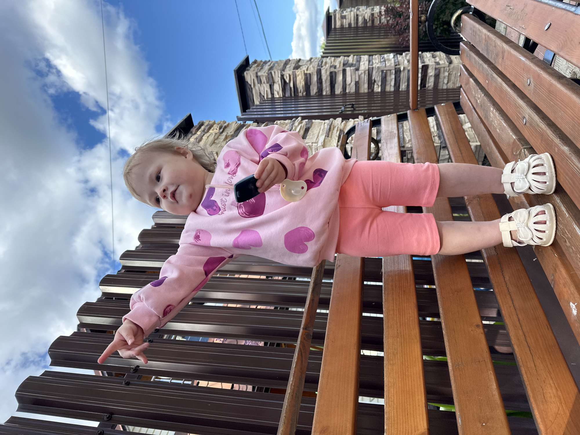 A young child, likely a toddler, is depicted lying horizontally on a wooden slatted structure, possibly a bench or swing, in an outdoor setting. The child has light-colored hair and is wearing a pink long-sleeved top adorned with darker pink heart patterns, pink pants, and white open-toed sandals.
The child's head is positioned on the right side of the frame, turned towards the left, with a curious or focused expression. Their right arm is extended towards the left side of the frame, with the index finger pointing downwards towards a dark brown corrugated or slatted metal fence visible in the lower left quadrant of the image. The child's left hand, positioned closer to their body on the right, holds a small black smartphone-like device and a yellow pacifier. Text "just in love" is faintly visible on the child's pink top.
The background features a bright blue sky with white clouds occupying the upper left portion of the image. In the upper right, behind the child, there is a building or wall with a textured, possibly stone or faux stone, finish. Some greenery is also visible beyond this structure. The bright natural lighting suggests it is daytime. The scene captures a casual moment in what appears to be a garden or yard in Nizhny Novgorod, Russia.