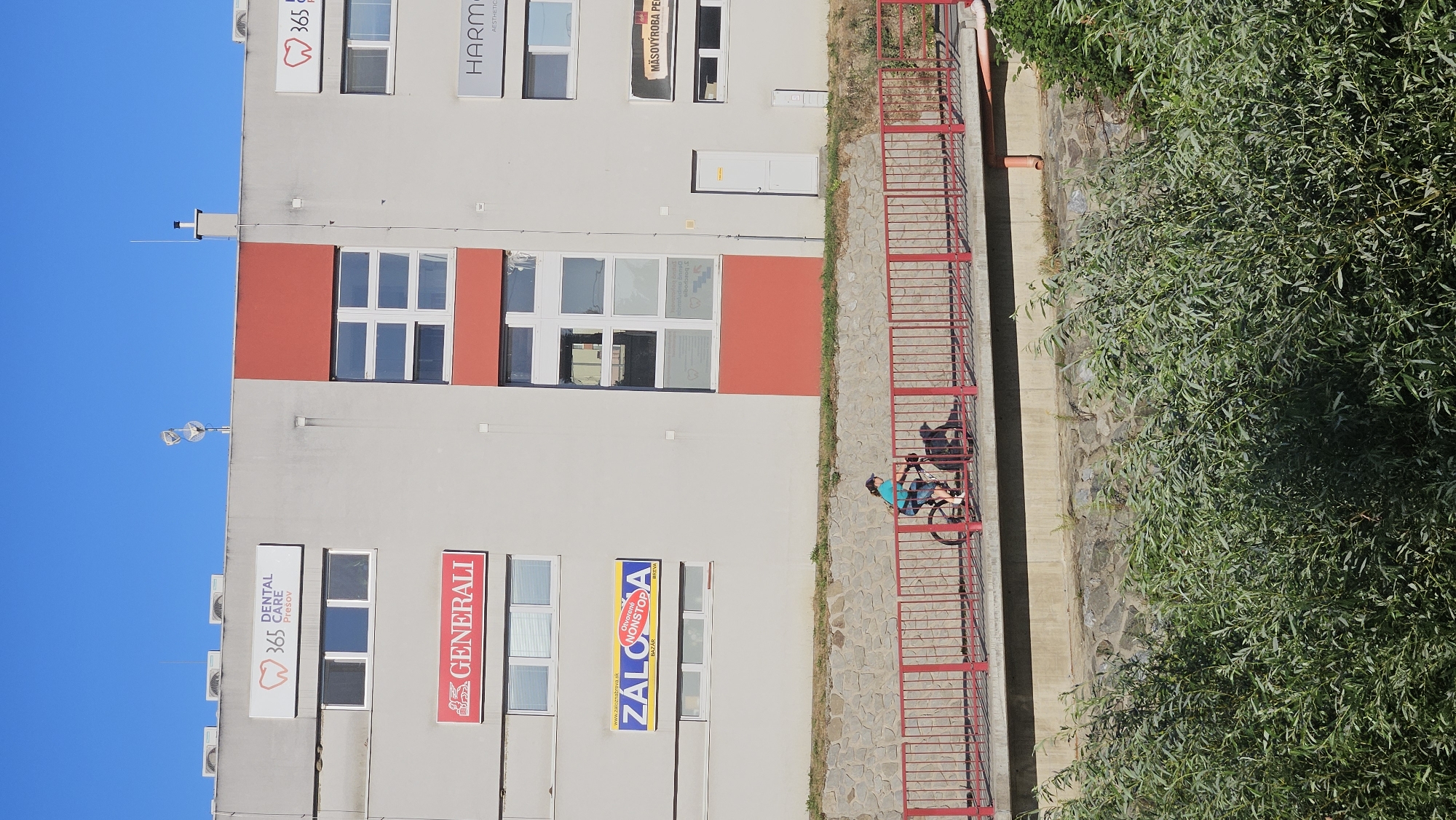 A young person on a bicycle rides across a red bridge that spans a small concrete-lined waterway. The bridge has a red railing. Behind the bridge, a three-story building stands with various businesses advertised on its facade. Signs visible include "365 DENTAL CARE Presov", "GENERALI", "ZALOZNA BAZAR NONSTOP", "HARMONY", and "MASOVYROBA". The building has multiple windows, some of which are tinted. The sky is a clear blue. Green foliage lines the bottom of the image in the foreground, obscuring the base of the building and the waterway. The overall scene depicts an urban environment with recreational activity.