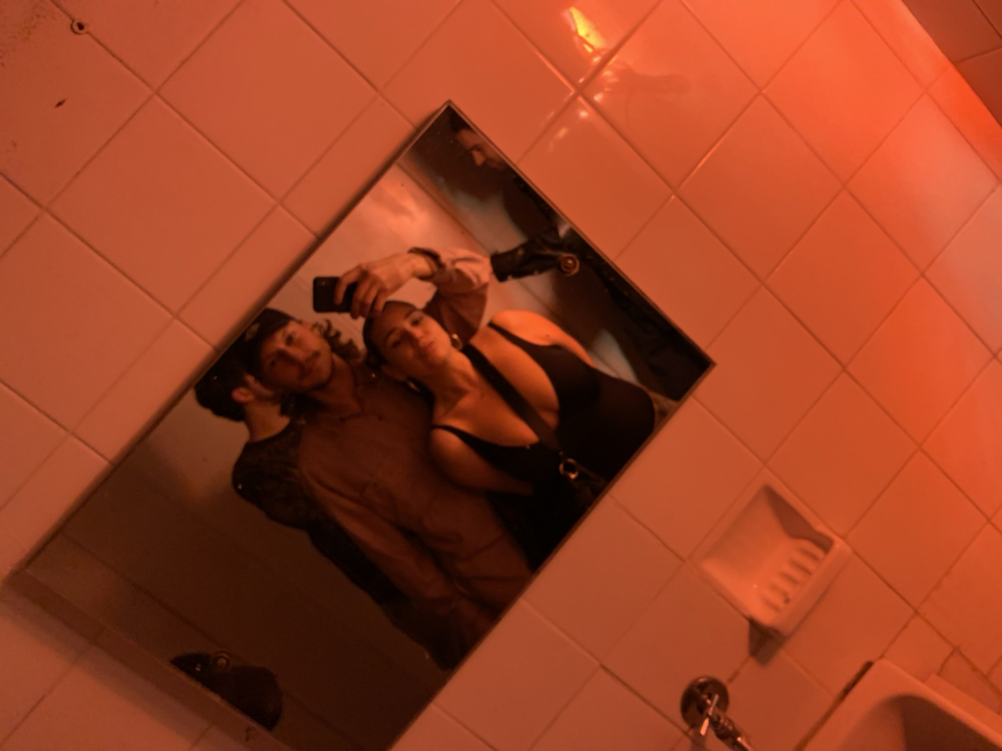 The image depicts a mirror selfie of two individuals, a male and a female, taken within a bathroom illuminated by a dominant red light source. The environment features light-colored square wall tiles with visible grout lines. In the lower-left corner, a partial view of a white sink or basin, a chrome faucet, and a recessed white ceramic soap dish are visible. The mirror, framed in dark material, reflects the subjects.
The female is positioned in the foreground of the reflection, holding a dark-colored smartphone in her raised left hand to capture the image. She is wearing a dark-colored tank top or bodysuit and has dark hair, potentially styled upward. Her facial expression is directed towards the camera. The male stands behind her and slightly to her right, with his right arm around her left shoulder. He wears a light-colored, long-sleeved collared shirt over a darker undershirt and a dark baseball cap worn backward. He has a mustache and visible dark hair extending from beneath his cap. Both individuals are posing for the camera.
Based on the provided information, this scene is situated within a public or semi-public restroom in the city of Tel-Aviv, Israel, likely a bar, club, or restaurant, given the aesthetic and context of the photograph.