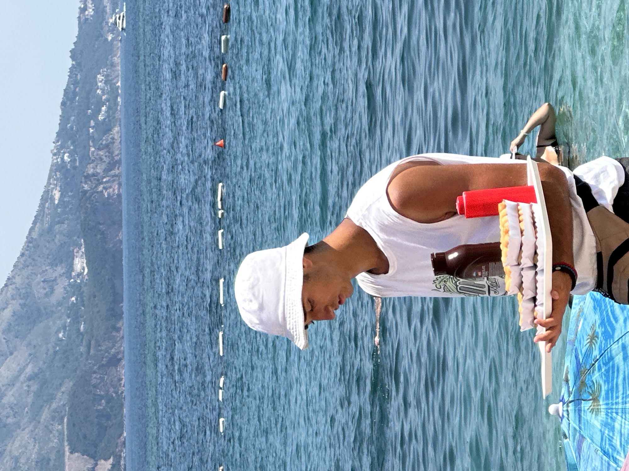 In Budva, Montenegro, a young man wearing a white bucket hat and a white t-shirt stands in the shallow blue water of the Adriatic Sea, holding a tray of food. The tray contains several white paper boats filled with what appear to be waffles, and a bottle of dark sauce, possibly chocolate syrup, and a red squeeze bottle, likely ketchup, are also on the tray. Behind the man, the sea stretches towards a lush green mountainside dotted with buildings, and a white yacht is visible in the distance. The scene suggests a busy beach day, with buoys marking a swimming area in the water and a blue beach umbrella partially visible in the foreground.