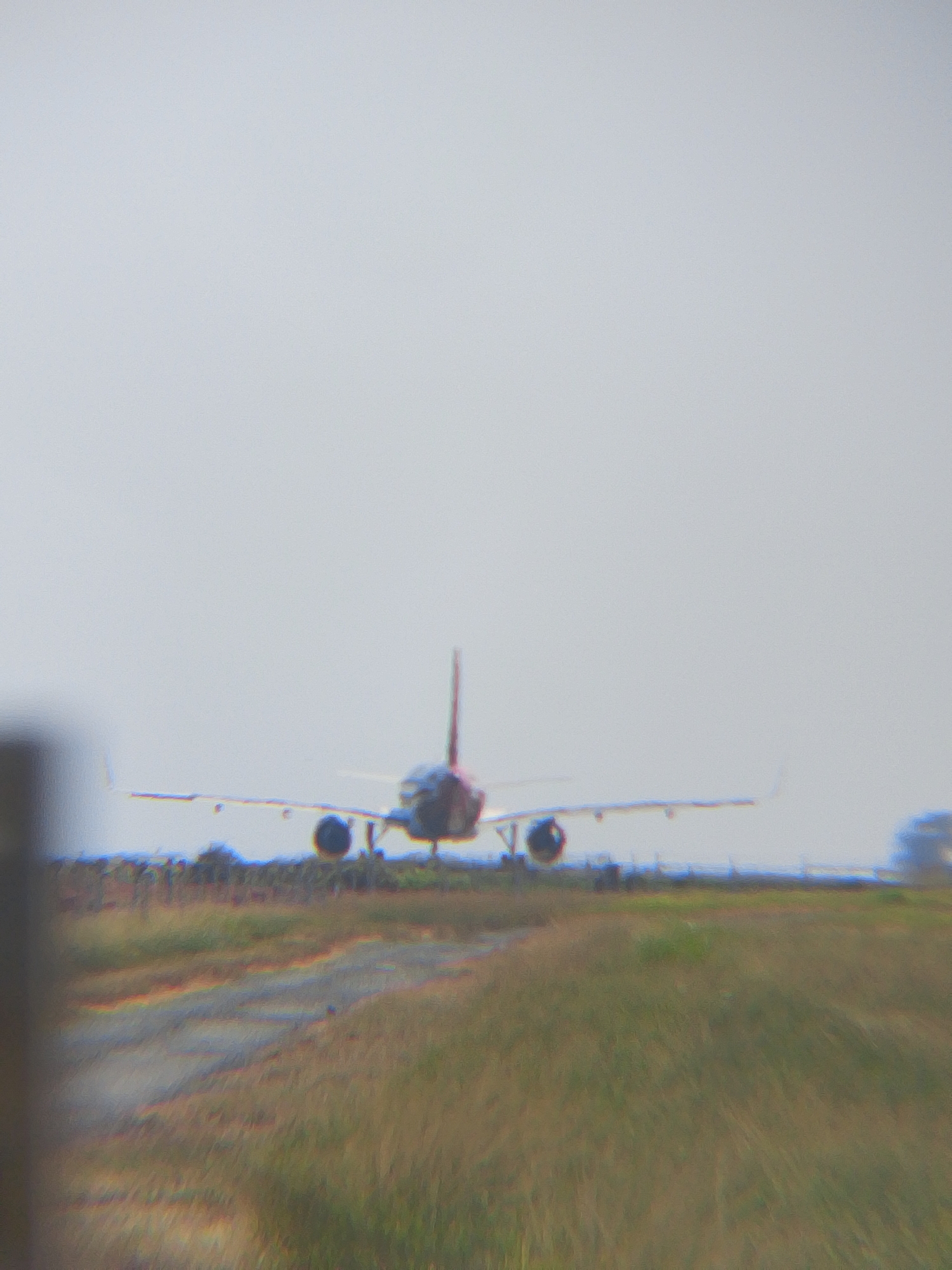 The image depicts a commercial airplane, seen from behind, on a runway with a blurred foreground and background. The sky is overcast and a pale gray. The airplane is mostly white with dark engines and a red and white tail. It is positioned in the center of the frame, facing away from the viewer, with its wings extended. The runway appears to be made of asphalt or concrete, and it is flanked by grassy areas. The overall impression is one of an outdoor scene, likely an airport, with a focus on the airplane. The image is out of focus, particularly in the foreground and background.
While this image depicts an airplane which could be in Balikpapan, Indonesia, the image itself does not contain specific visual information that would allow for a definitive identification of the location within Balikpapan. The general setting is consistent with an airport environment.