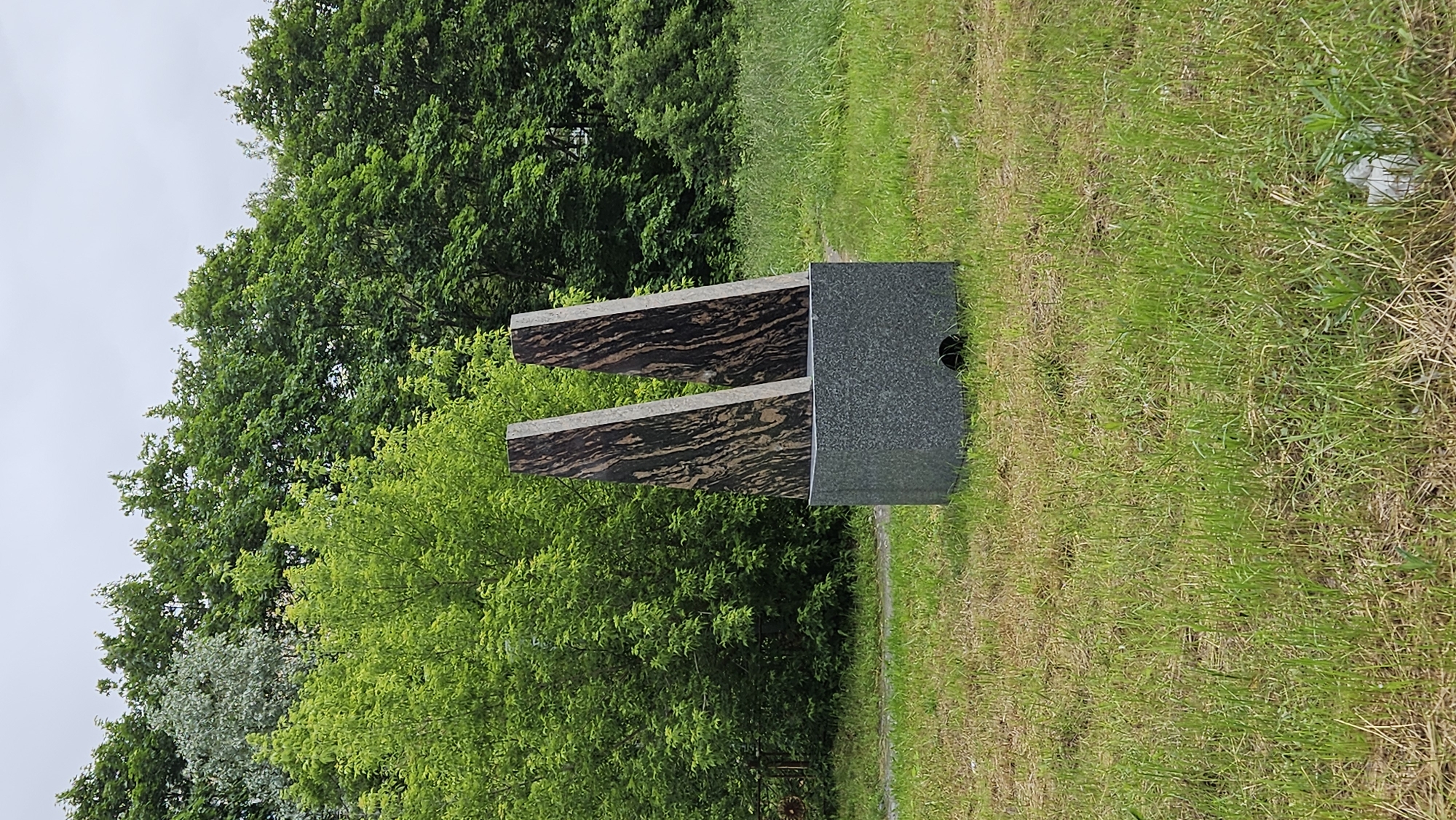 An outdoor, eye-level, medium shot depicts a monument in a grassy field. The monument consists of a gray, rectangular base with two angled, dark brown and tan stone slabs rising from its center. Both slabs are bordered with a lighter tan stone. The base has a circular opening on its lower right side. The field is covered in green and brown grass, with some dry patches. In the background, a dense line of green trees forms a natural backdrop under a cloudy sky. The overall environment appears natural and unkempt.