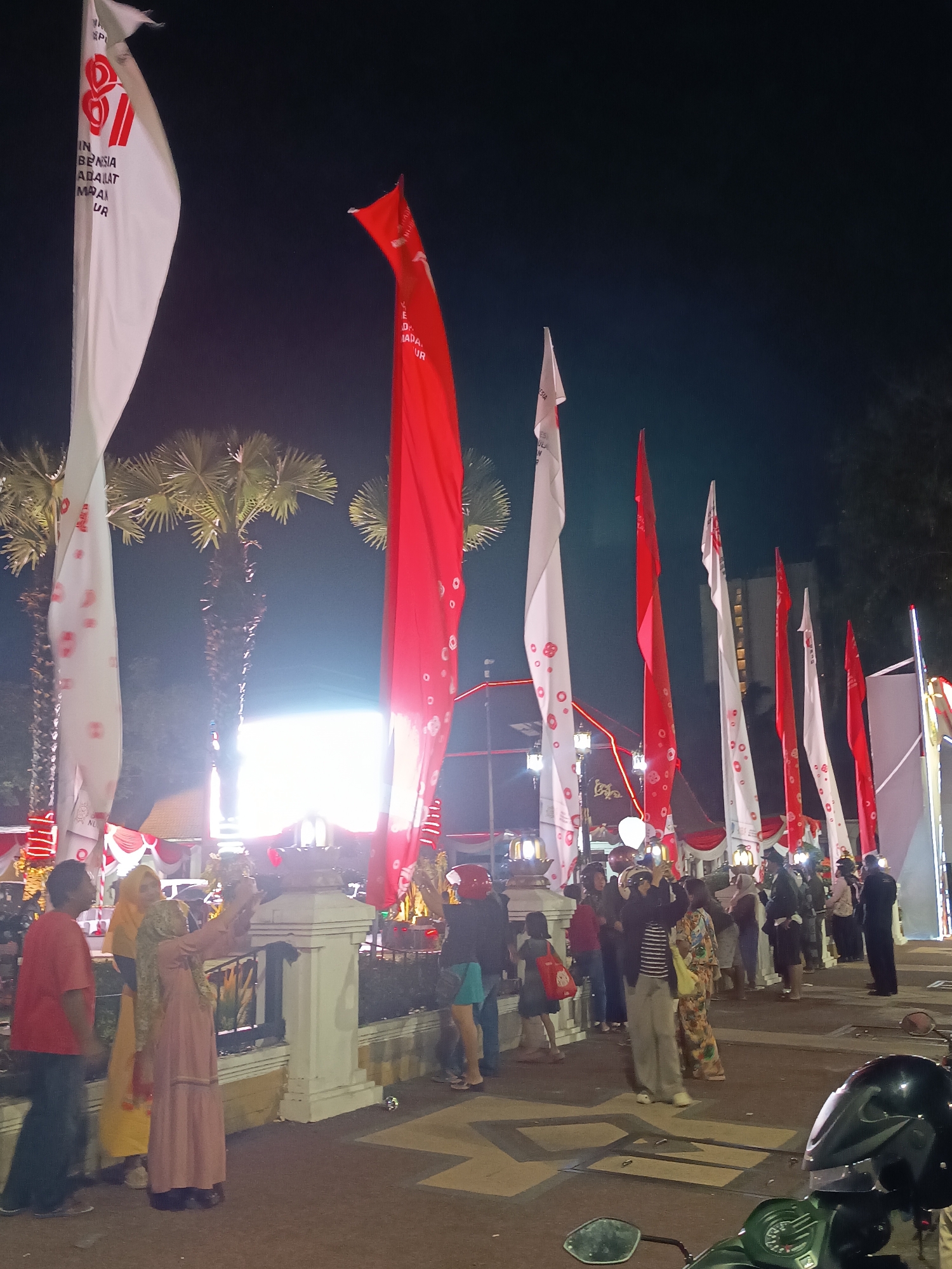 The image depicts an outdoor nighttime scene, likely in a public space within the city of Surabaya, Indonesia, suggested by the prominent red and white flags which are characteristic of Indonesian national celebrations. The foreground shows a paved area with several people standing and walking. On the left, a man in a red t-shirt and dark pants is walking away from the camera. Beside him, a woman in a yellow hijab and a woman in a light pink dress are taking a photo with a smartphone. Behind them, a white pillar structure is visible. A line of tall, red and white flags, some bearing circular patterns, are erected on poles and are a dominant feature of the scene, stretching across the midground and into the background. Palm trees with visible fronds are scattered amongst the flags. In the background, there appears to be a brightly lit structure, possibly a building or stage, with decorative lighting. Further back, other people are milling around, some dressed in formal or semi-formal attire, and a motorcycle is parked in the lower right corner of the frame, partially obscuring the view. The overall atmosphere suggests a festive event or gathering.