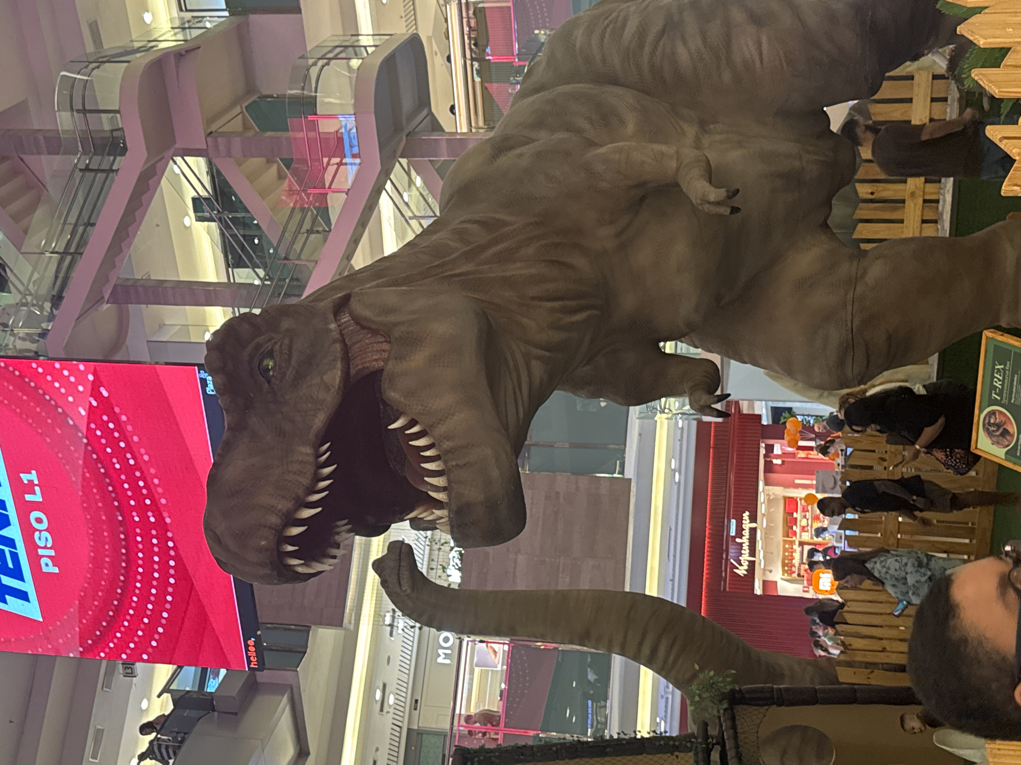 A large, realistic Tyrannosaurus Rex sculpture is the central focus of this indoor scene, possibly at a shopping mall in Niterói, Brazil. The dinosaur is depicted with its mouth open, revealing sharp teeth and a reddish interior, and its head is turned to the right. To the left of the T-Rex, a long, tan neck and head of another dinosaur, possibly a Brontosaurus, is visible. The background features elements of a shopping mall, including escalators with pink railings, shops with signage like "Kopenhagen" and "M", and various people walking around or observing the display. A large red digital screen displaying "TENSO PISO L1" is prominently situated above and to the left of the T-Rex. The lighting appears to be artificial, common in indoor commercial spaces.