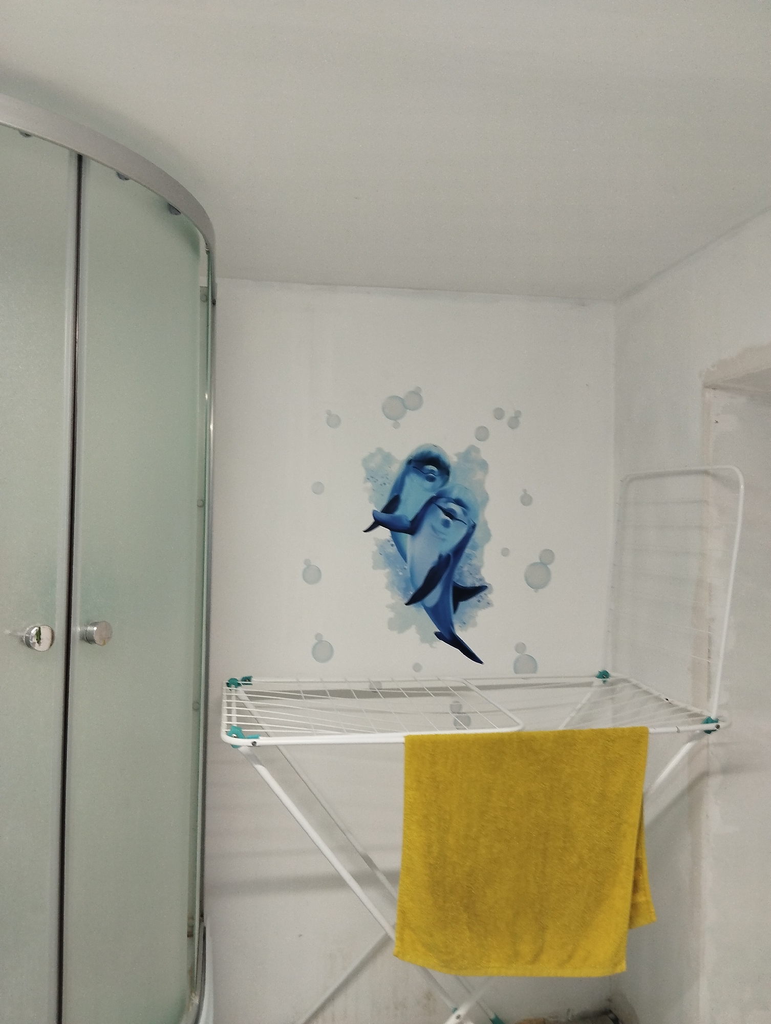The image shows a bathroom in Krasnogorskoye, Russia. A rounded frosted glass shower stall is to the left. On the wall is a blue dolphin decal with white bubble accents. A white folding drying rack is placed in front of the wall with a folded yellow towel hanging on it. The walls and ceiling are white.