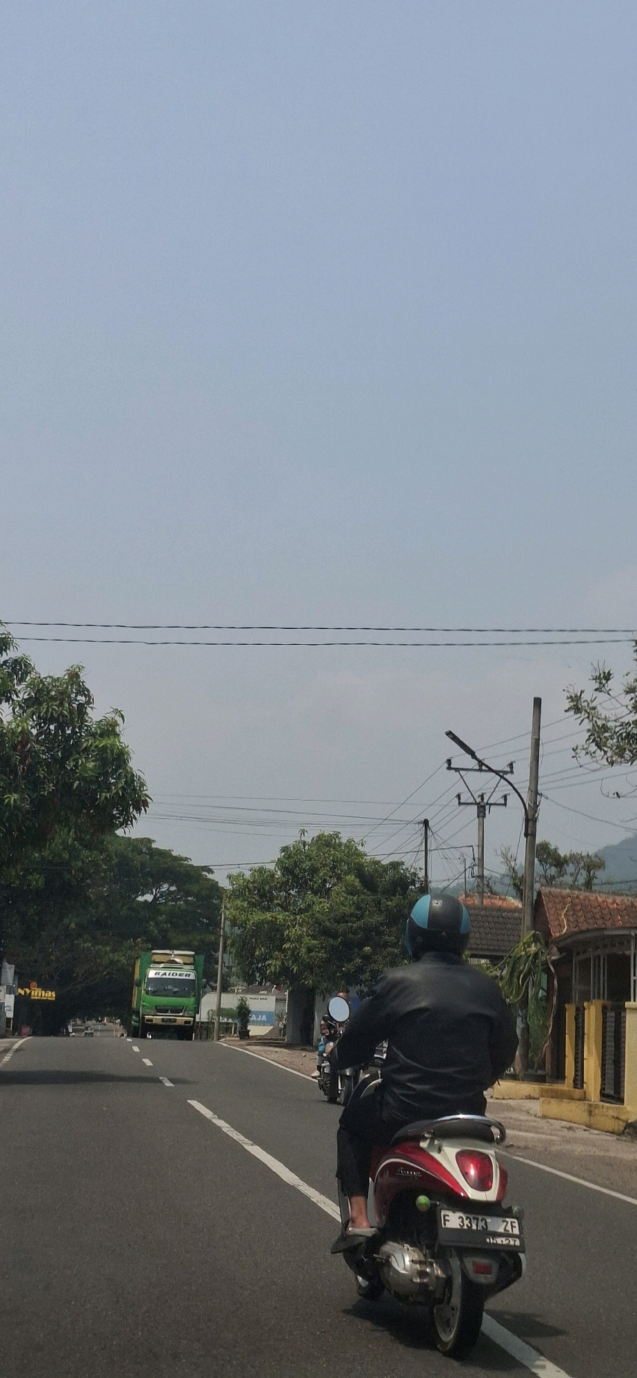 In Susukan, Indonesia, a person wearing a black jacket and a blue and black helmet is riding a red and white scooter on a paved road. The scooter has a license plate that reads "F 3373 ZF". The person's feet are visible, wearing grey sandals. The scooter has a green object attached to its rear. The road is marked with white lines. In the background, a green truck with the word "RAIDER" on its side is visible. Several trees line the side of the road, and some buildings with tiled roofs are also present. Overhead, power lines are strung across the sky. The sky is a pale blue with some haze. Another person on a scooter is visible further down the road, in front of the truck. The scene depicts a typical daytime street view in a semi-urban or rural Indonesian setting.