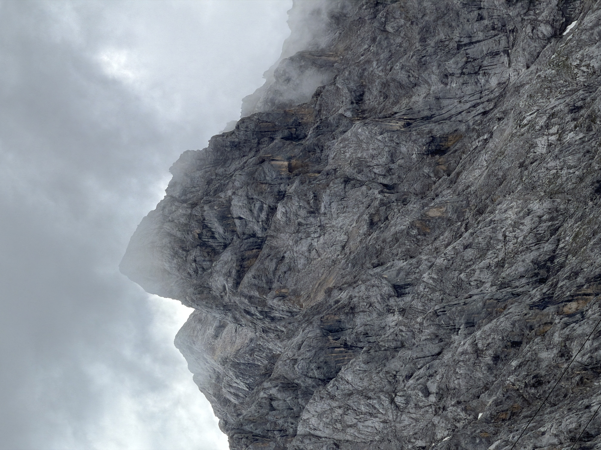 The image displays a dramatic, close-up view of a jagged, rocky mountain face on a cloudy day. The mountain's surface is heavily textured with numerous fissures, ridges, and sheer drops, rendered in shades of grey and dark grey. Patches of mist cling to the upper portions of the mountain and the sky above is overcast with heavy, textured grey clouds. The overall impression is one of rugged, imposing natural terrain under a moody, atmospheric sky. The mountain is likely Zugspitze, Germany's highest peak, located in the Wetterstein range near Garmisch-Partenkirchen. There are no people, animals, or man-made objects clearly visible in the frame, apart from what might be thin wires or cables running diagonally across the lower portion of the mountain face, potentially part of a cable car system.