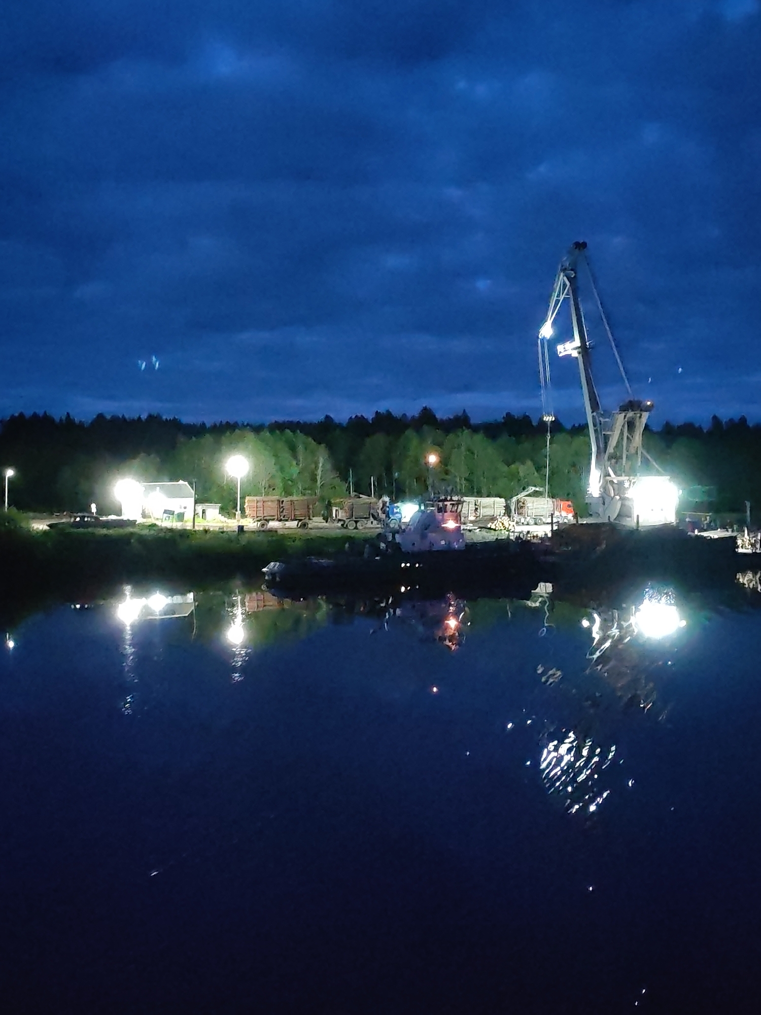 The image shows a nighttime scene at Annensky Most, Russia. A body of water reflects the lights from the industrial area along the shoreline. A tall crane is prominent, illuminated by lights. Several trucks are parked near the crane, and a small white building is illuminated by a light near the left edge. Trees line the horizon. The sky is dark, with some visible clouds.