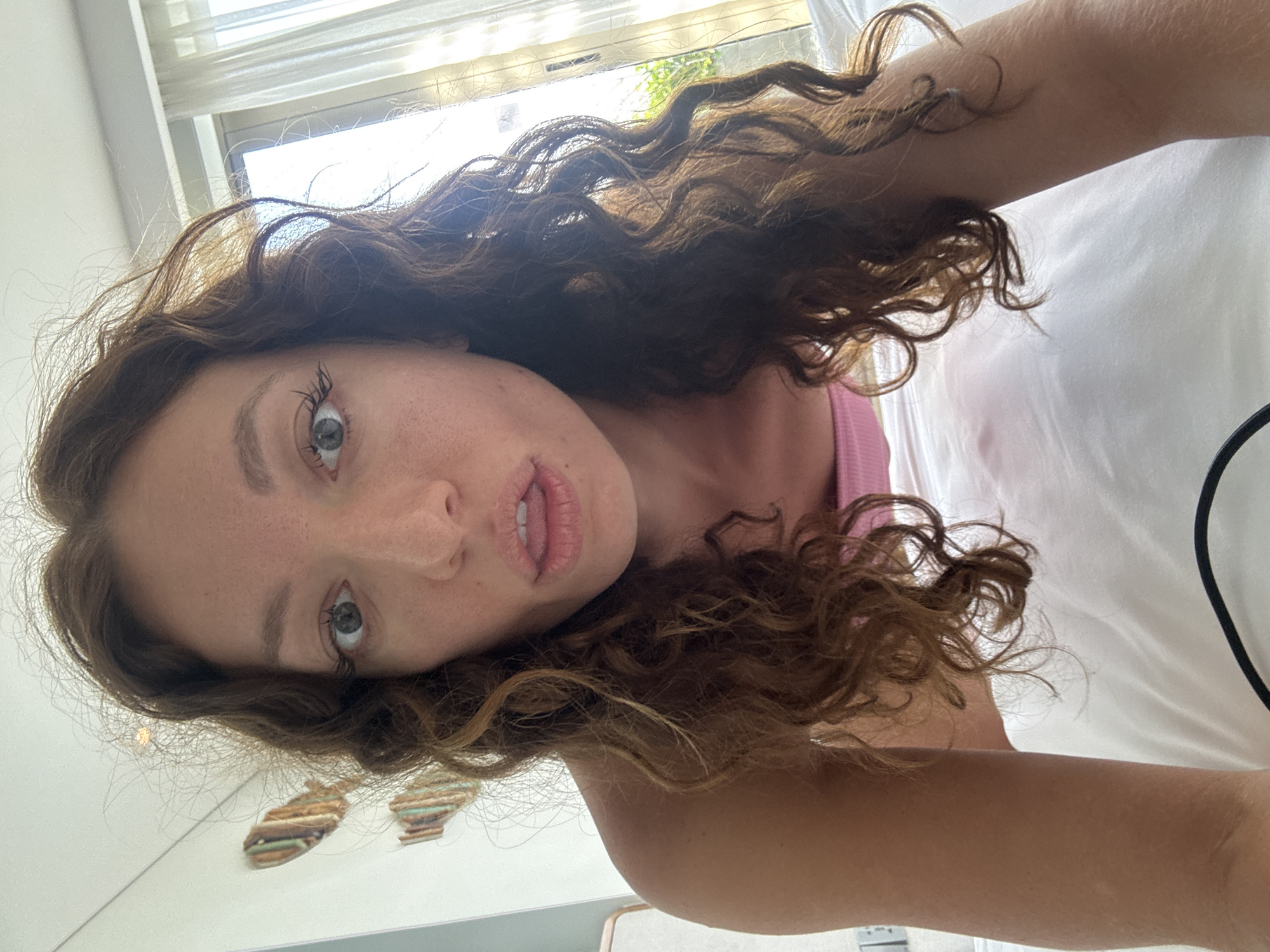 A woman with curly brown hair and fair skin is lying on a white bed. She has gray eyes and a slightly parted mouth. She is wearing a pink top. A window with light filtering through the blinds is visible in the background. There is no evidence to suggest that the image was taken in Đà Nẵng, Vietnam.