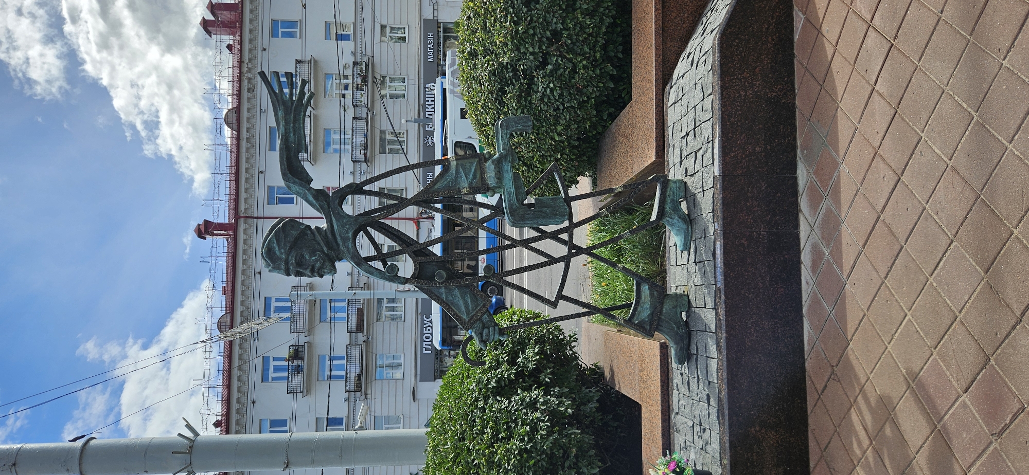 In Vitsebsk, Belarus, this photograph captures a distinctive bronze sculpture of a man walking, situated on a paved plaza. The sculpture depicts a figure with an exposed, skeletal-like frame, wearing a hat and a scarf that billows outwards, suggesting movement. He carries a cane in his left hand and appears to be holding a rectangular object with his right. The sculpture stands on a base constructed from granite and stone pavers laid in a diamond pattern. Behind the sculpture, a white building with red-tiled roofs and balconies dominates the background. Trees and bushes flank the sculpture on either side, and a blue bus with the word "ГЛОБУС" (Globus) visible on its side drives past in the mid-ground, with other signs, including "БИБЛИОТЕКА" (Library) and "МАГАЗИН" (Shop), also discernible on the building facade. The sky above is blue with scattered white clouds, and a utility pole with wires is visible on the left side of the frame.