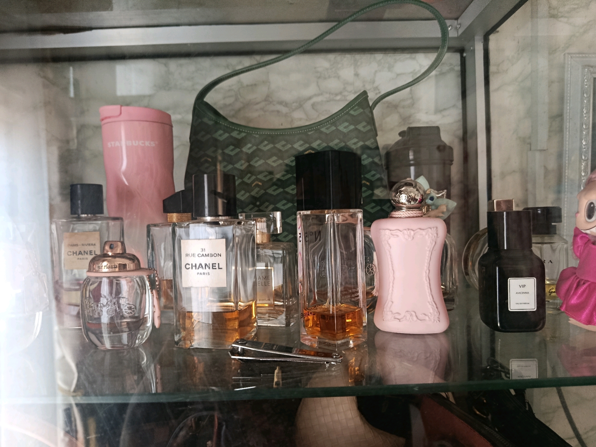 The image shows a shelf in a display case with various perfume bottles, a pink Starbucks tumbler, and a green designer handbag. The perfumes are arranged in rows, with some bottles larger and others smaller. The Starbucks tumbler is pink and has "STARBUCKS" written on it in white. The handbag is green and has a geometric pattern. A small, decorative bottle with a silver top and a light blue charm is also visible. In the background, there are other objects, including what appears to be a framed picture and a doll-like figurine in a pink outfit. The lighting is somewhat dim, with reflections visible on the glass of the display case. The overall scene suggests a personal collection of fragrances and accessories.