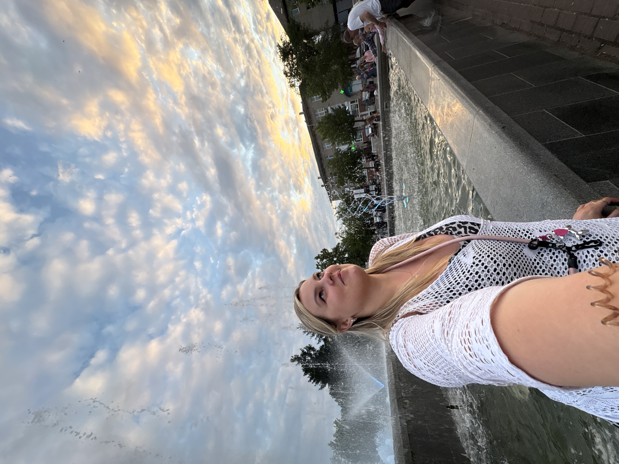 An outdoor, eye-level, selfie-style photograph captures a blonde woman standing beside a public fountain at dusk, with a dramatic sky overhead. The woman is wearing a white, open-knit cover-up over a black and white patterned swimsuit. Her head is tilted upwards, and she is looking to her right, with a slight smile. She is holding the camera with her left hand, which is visible in the foreground. The fountain features several jets of water, some illuminated with blue light. In the background, there are trees, buildings, and other people near the fountain, suggesting a public square or park. The sky is filled with dynamic clouds, illuminated by the setting sun, casting warm hues of orange and yellow. This scene is located in Chaikovsky, Russia.