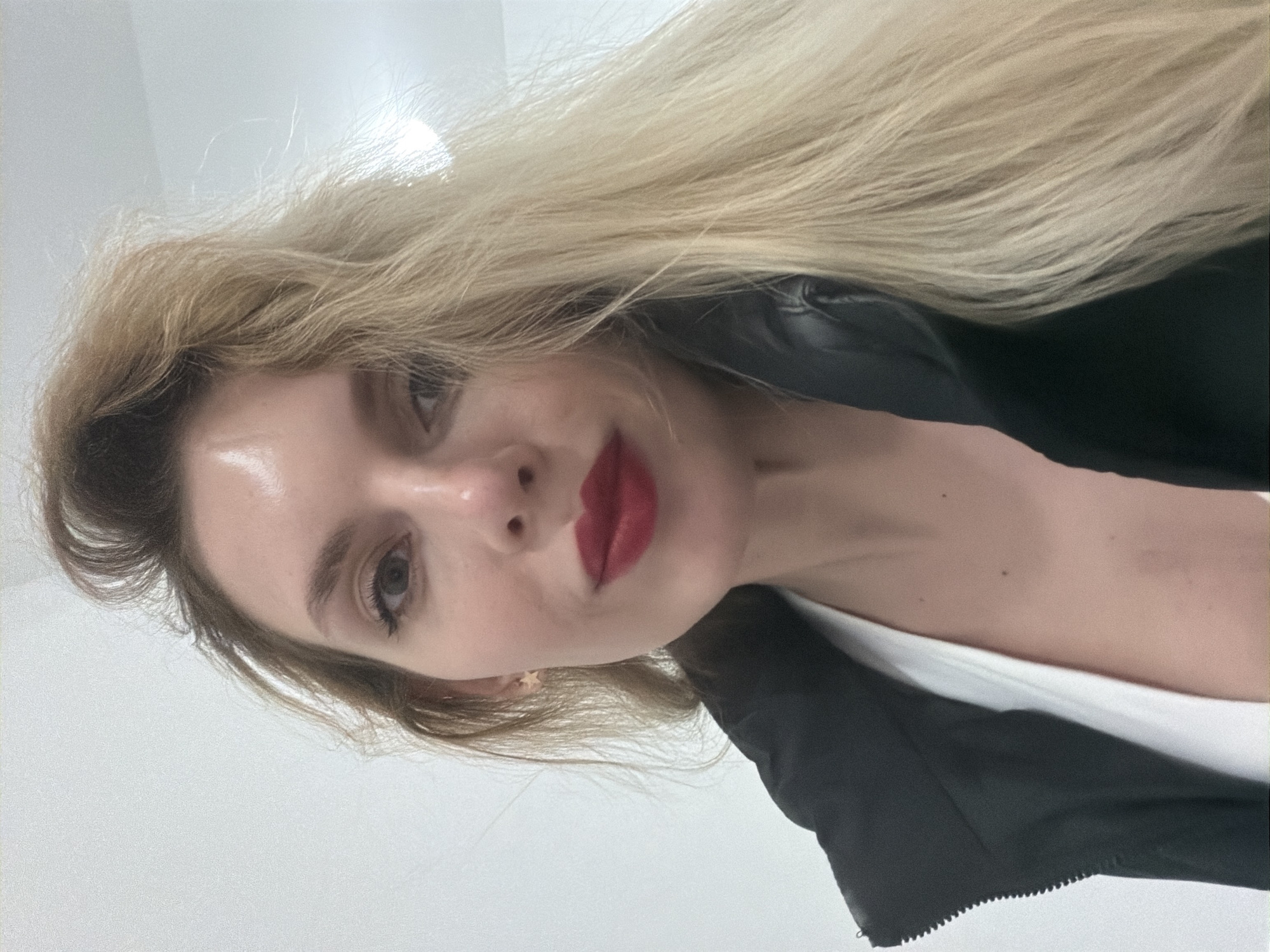 This close-up portrait features a woman with long, straight blonde hair cascading across the frame, flowing from the upper left towards the lower right. Her head is tilted, positioned on the left side of the image, with her body extending diagonally downwards. She has fair skin, prominent red lipstick, and dark eyelashes framing her light-colored eyes. Her gaze is directed towards the viewer, accompanied by a subtle smile.
She is wearing a black outerwear garment, possibly a jacket, which has a visible zipper detail along the bottom edge of the frame. A white top is visible underneath the black layer. A small, golden, star-shaped earring adorns her left earlobe. The background is a plain, bright, off-white or very light grey, suggesting an indoor setting with minimal distractions. The image is brightly and evenly lit. While no specific location cues are visible within the image, the provided context indicates Budva, Montenegro. There is no visible text.