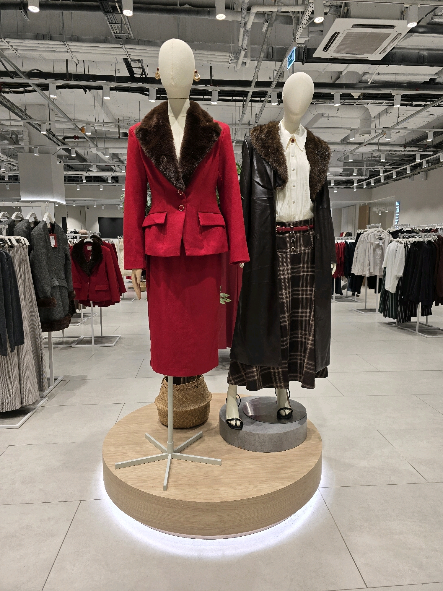 This image shows two mannequins displayed in a retail store in Khimki, Russia. The mannequins are positioned on a circular wooden platform with LED lighting at its base. The mannequin on the left is wearing a red suit, consisting of a jacket with a fur collar and a matching skirt. The mannequin on the right is dressed in a white blouse, a dark long coat with a fur collar, and a plaid skirt with a red belt. Behind the mannequins, racks of clothing are visible, suggesting this is a clothing store. The ceiling is exposed, revealing various pipes, electrical conduits, and lighting fixtures, characteristic of a modern retail environment. The floor is tiled with large, light-colored tiles.