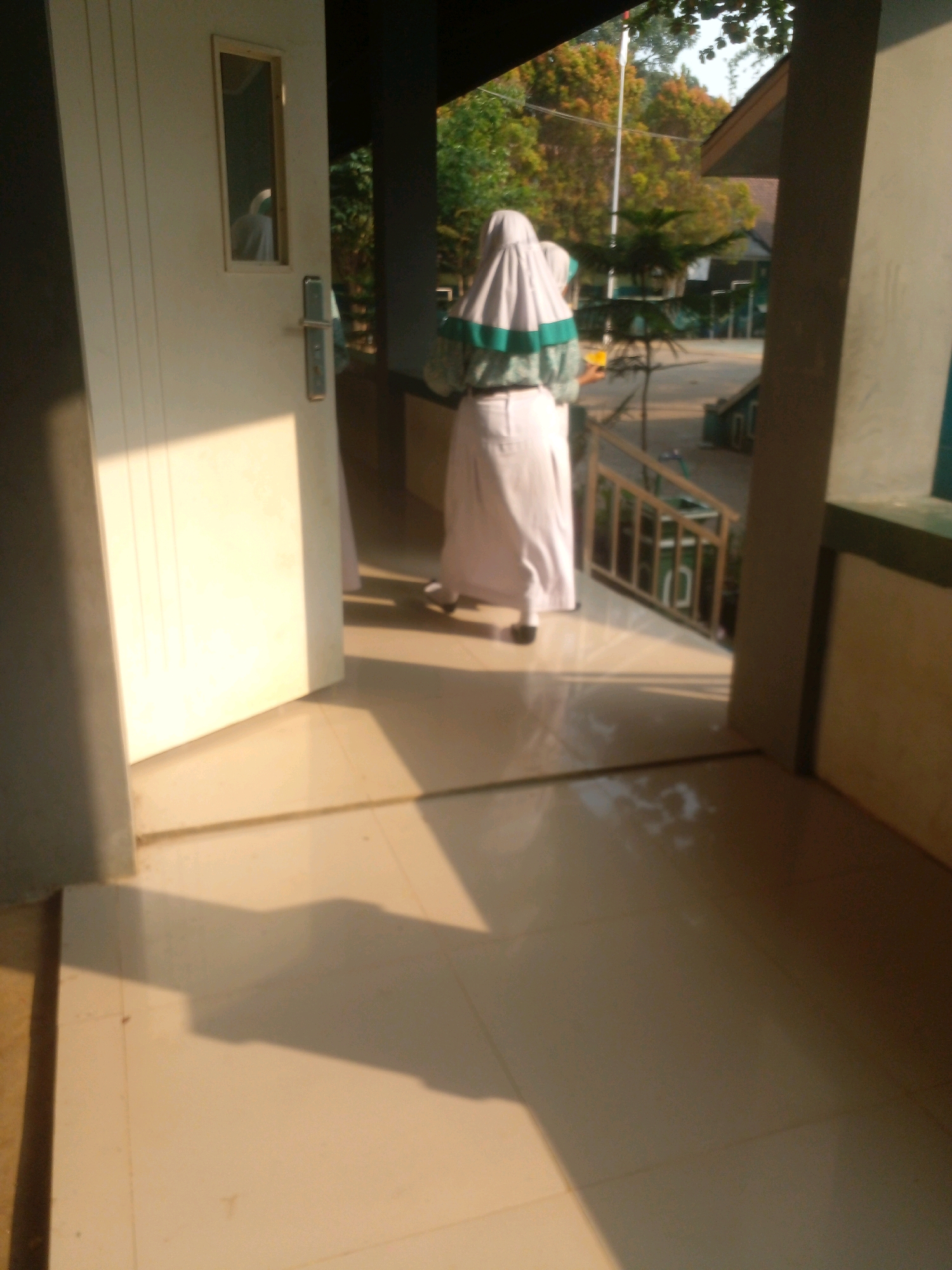A young girl in a hijab and a long skirt walks away from a white door in an outdoor corridor of a school in Banyuputih, Indonesia. The girl's hijab is gray with green stripes. She is holding a bright orange cup in her left hand. The corridor has a tiled floor and a railing along the side. The sun casts long shadows across the floor. In the background, there are trees and buildings, suggesting an educational institution.