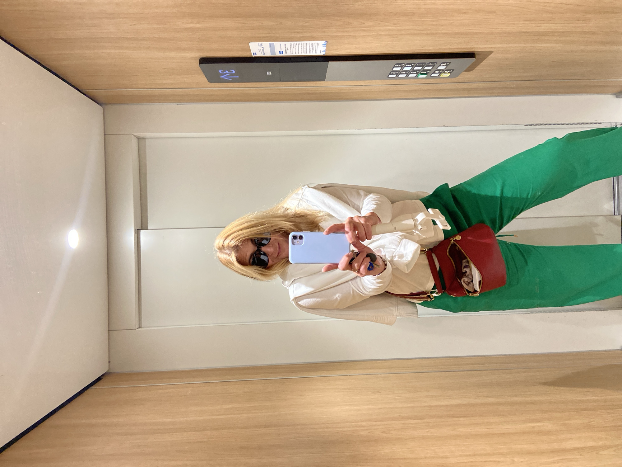 A woman wearing sunglasses, a white top, green pants, and a red purse takes a selfie with a blue smartphone inside an elevator. The elevator has a wood panel interior, a control panel with buttons and a digital floor display showing "3V", and a card with writing above the control panel. The walls appear to be off-white. The location is possibly in Budva, Montenegro.