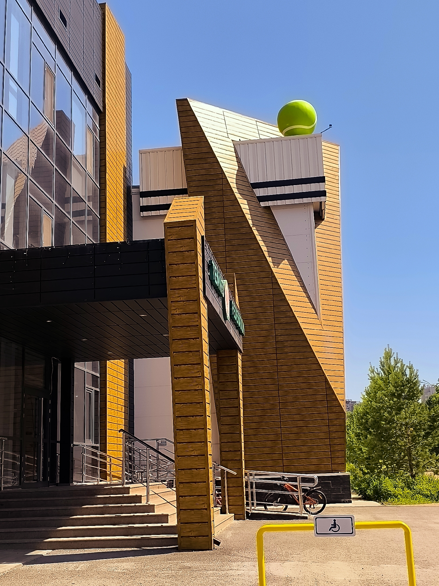 A modern building with a glass facade and wooden paneling is seen under a clear blue sky. A giant green tennis ball rests atop the angled roofline of a section of the building. In front of the entrance, a set of concrete stairs leads up to a covered walkway. A bicycle is parked near a ramp, and a yellow barrier with a wheelchair accessibility symbol is visible. The scene is set in an urban environment, likely in the city of Qarağandı, Kazakhstan, as suggested by the context.