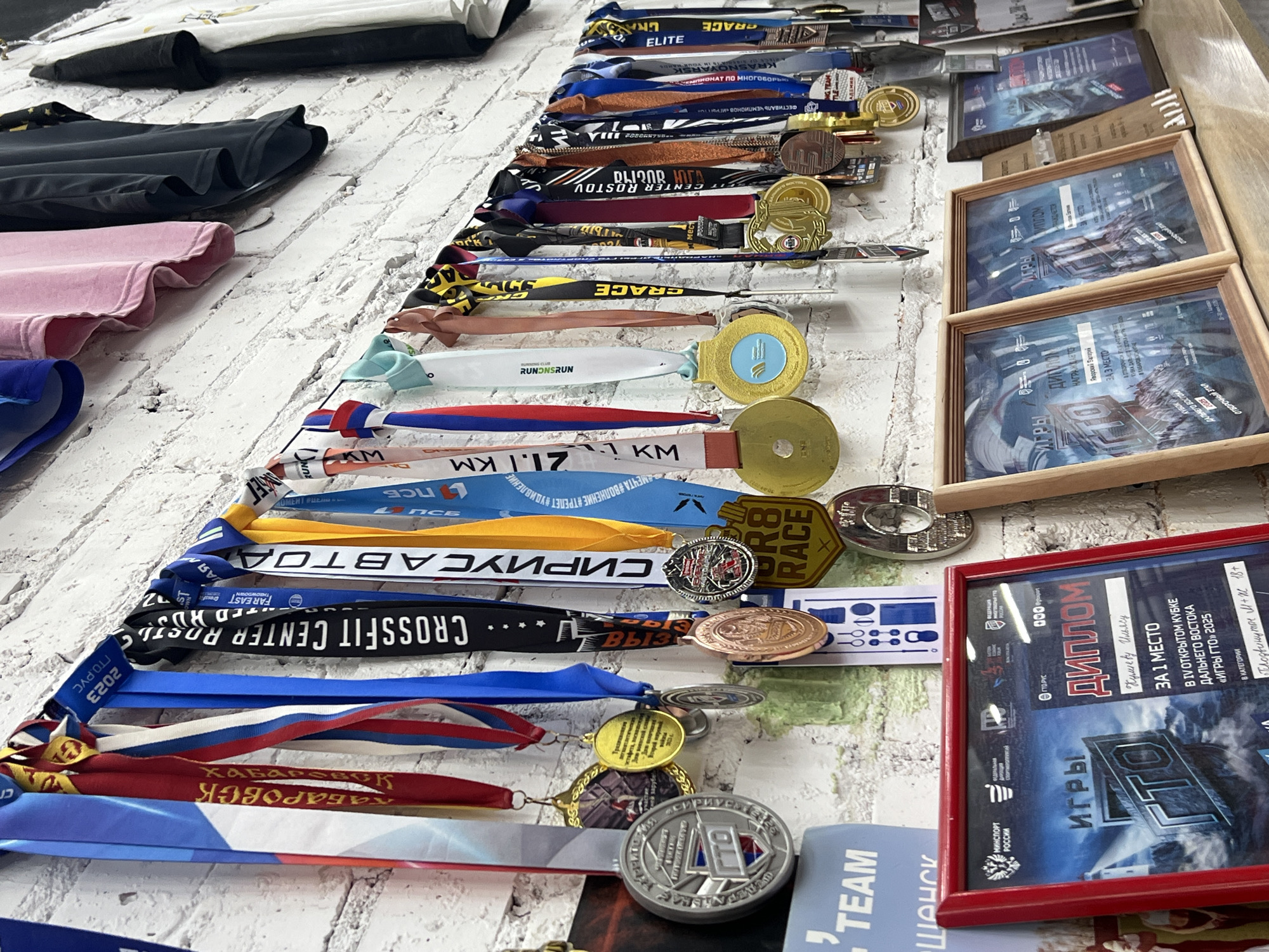 The image displays a proud collection of numerous sports medals and framed diplomas, meticulously arranged on a white, brick-patterned surface, likely indoors. To the left, folded athletic wear in black, pink, and blue is stacked.
The primary subjects are the extensive array of medals and awards. The medals are diverse, featuring circular metallic designs in gold, silver, and bronze hues, alongside some uniquely shaped pieces like the "R8 RACE" medal. These are attached to a variety of colorful ribbons, many bearing inscriptions such as "GRACE," "RUNONSRUN," "CROSSFIT CENTER ROSTOV," "21.1 KM" (likely a half-marathon distance), and "ГТО РУС 2023." One prominent ribbon specifically names "Хабаровск" (Khabarovsk), providing a key location cue.
The framed diplomas, predominantly in Russian, attest to significant accomplishments. A red-framed diploma clearly awards "Кулиеву Ильясу" (Ilya Kuliev) "ЗА 1 МЕСТО" (for 1st Place) in the "IV ОТКРЫТОМ КУБКЕ ДАЛЬНЕГО ВОСТОКА «ИГРЫ ГТО» 2025" (IV Open Cup of the Far East "GTO Games" 2025) in the "Продвинутые М+Ж 18+" (Advanced M+W 18+) category. Other visible texts on ribbons and diplomas include "МИНСПОРТ РОССИИ" (Ministry of Sport of Russia), "ЧЕМПИОНАТ ПО МНОГОБОРЬЮ" (Championship in multi-event), and "ФЕСТИВАЛЬ ЧЕМПИОНОВ «ИГРЫ ГТО»."
The overall scene strongly suggests a display of athletic achievement and dedication, reflecting success in various sports competitions and physical fitness programs, particularly within the Far East region of Russia. The collection evokes a sense of pride and a history of competitive participation.