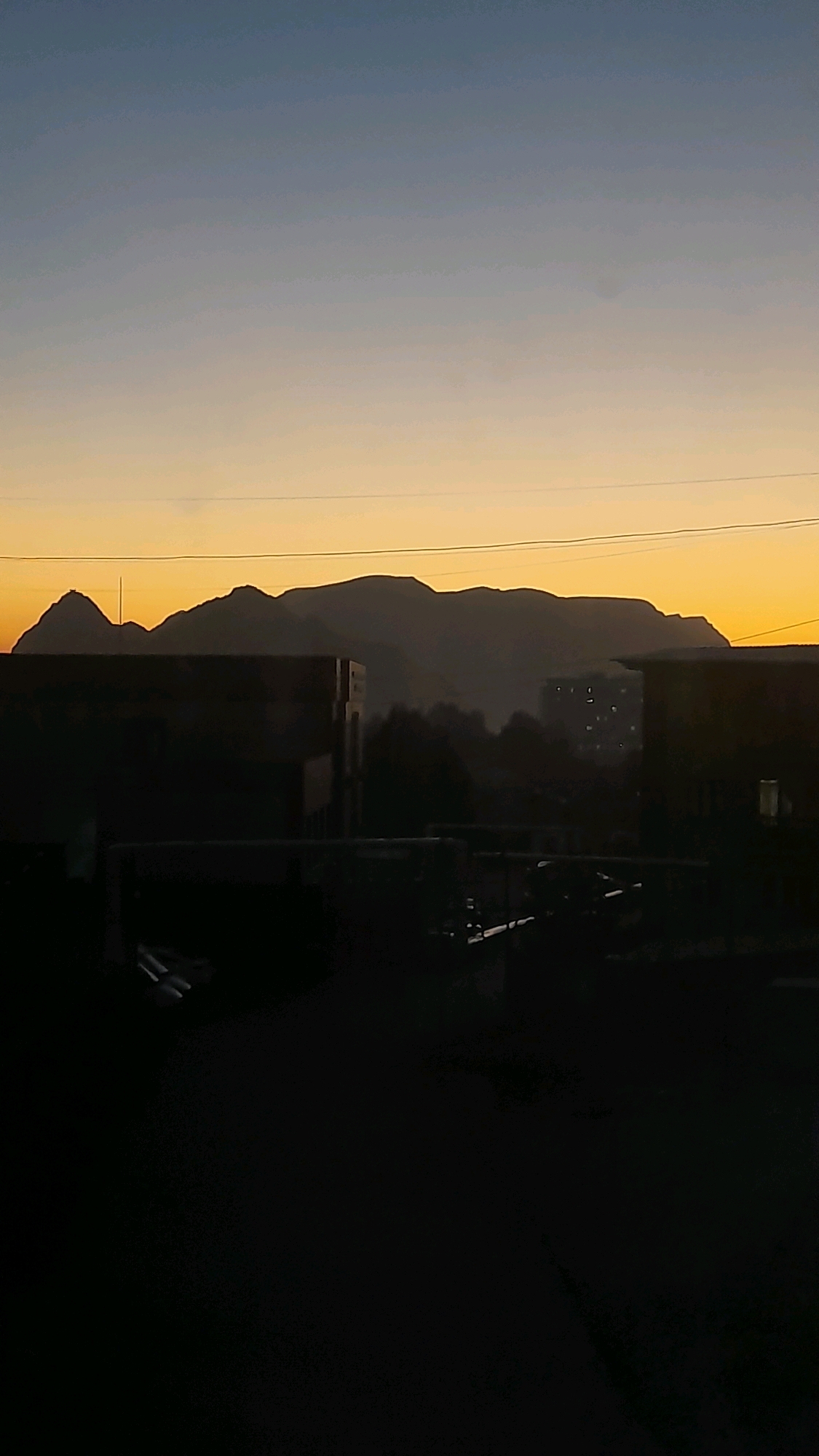 A panoramic photograph captures the silhouette of mountains against a gradient sky transitioning from deep blue at the top to a warm yellow-orange near the horizon. In the foreground, dark, indistinct shapes suggest buildings and structures, possibly in the city of Osh, Kyrgyzstan, with a few scattered lights visible in a distant building. Power lines stretch horizontally across the frame, dividing the sky. The overall impression is one of a twilight or dawn scene, with the natural landscape of mountains juxtaposed against the urban environment.