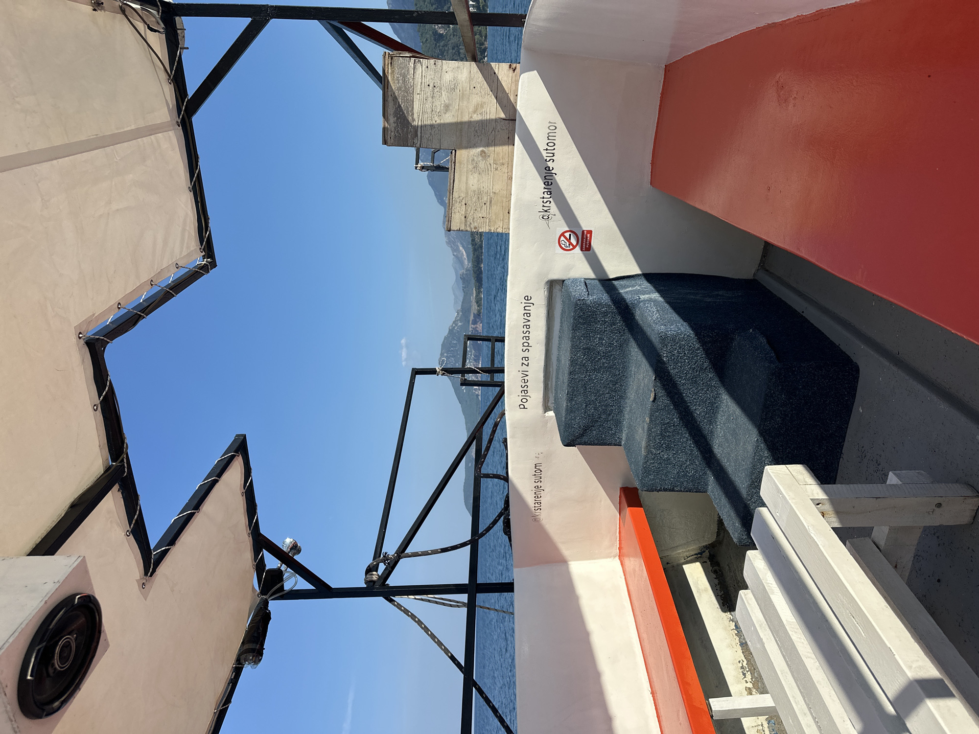 The image shows the interior of a boat at sea, with the coastline of Šušanj, Montenegro, visible in the distance under a clear blue sky. The boat has white and orange benches along the sides, and a dark blue carpeted staircase in the center. There are two signs on the white wall. One says "Pojasevi za spasavanje" which translates to "life jackets". The other sign, partially obscured by the staircase, has a red circle with a cigarette crossed out and text below that reads "ZABRANJENO PUSENJE", meaning "no smoking". A speaker is mounted on the overhead canopy. The water is calm and blue, and the coastline features green hills and some buildings. There are no people visible in the image.