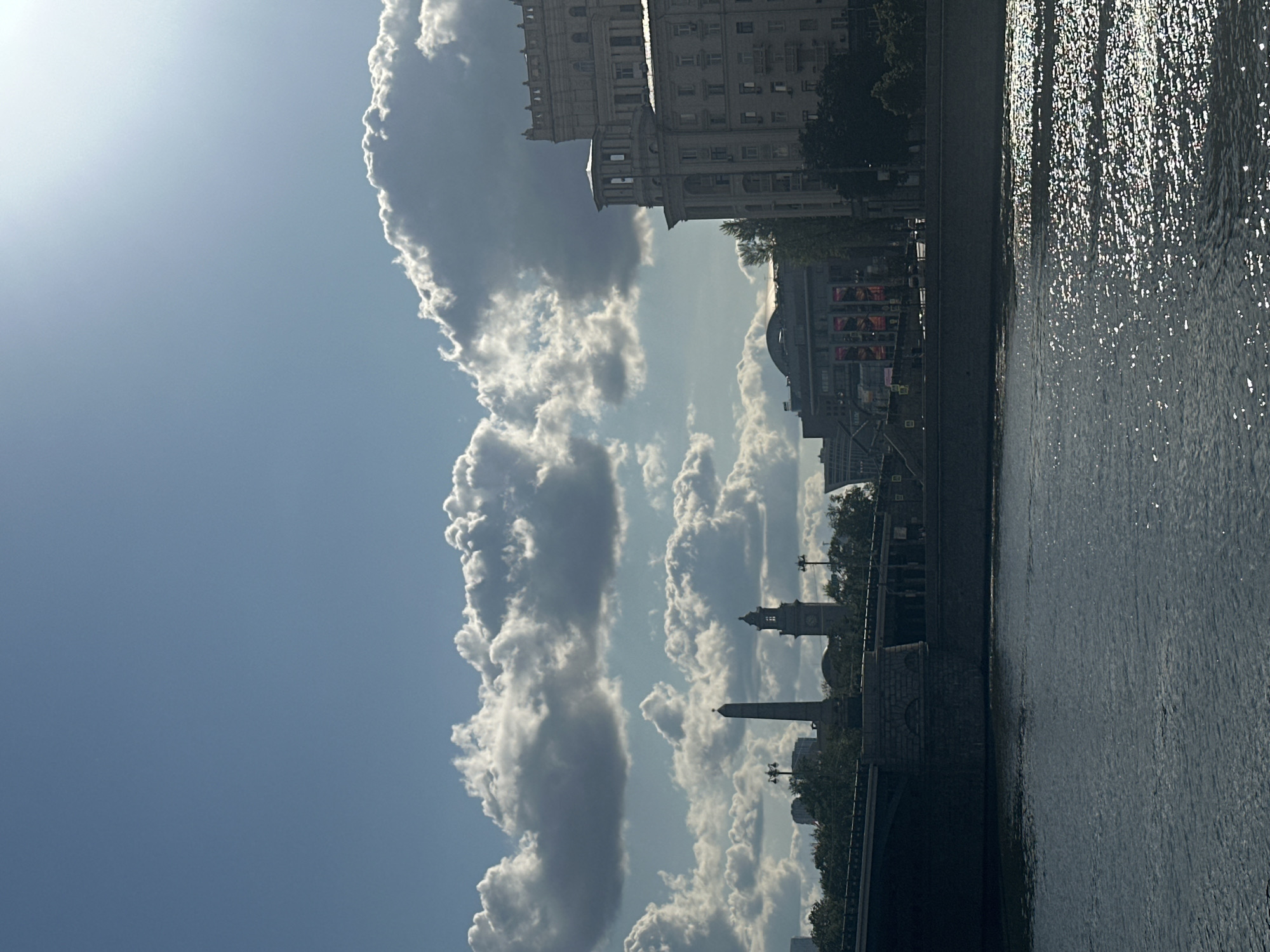 The image depicts a view of the Moskva River with buildings and landmarks in the background. The river is dark blue with sunlight glinting off its surface, suggesting a bright, possibly late afternoon, day. A stone bridge spans the river in the foreground, with a stone embankment lining the river on the far side.
In the background, several buildings are visible. To the left, an obelisk stands tall, and next to it is a clock tower. Further to the right, a large, ornate building with classical architectural elements dominates the skyline. Behind these, other urban structures and trees are discernible. The sky is a bright blue with scattered, fluffy white clouds, some of which are illuminated by the sun.
The scene appears to be located in Moscow, Russia, given the presence of landmarks like the obelisk and clock tower, which resemble structures found in the city, potentially near the area of the Moscow State University or the Novodevichy Convent. There are no people visible in the image. The overall impression is one of a sunny day in a city, with the river providing a prominent natural element.