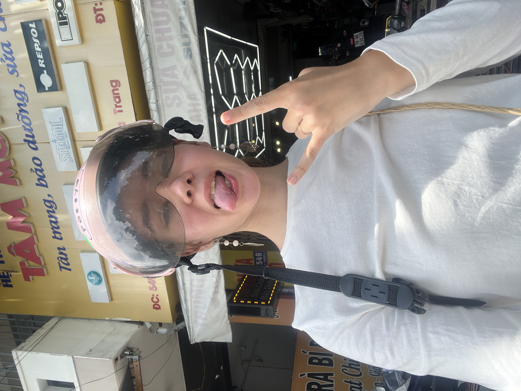 A young woman wearing a pink helmet with a clear visor is sticking her tongue out and making a rock and roll hand gesture in front of a shop in Nha Trang Ward, Vietnam. The shop has signs in Vietnamese, advertising motorcycle repair and supplies, including brands like "REPSOL," "SPEEDOL," and "SUZUKI." The street address "549" is visible on one sign. The woman is wearing a white, long-sleeved shirt, and a tan strap crosses her chest.