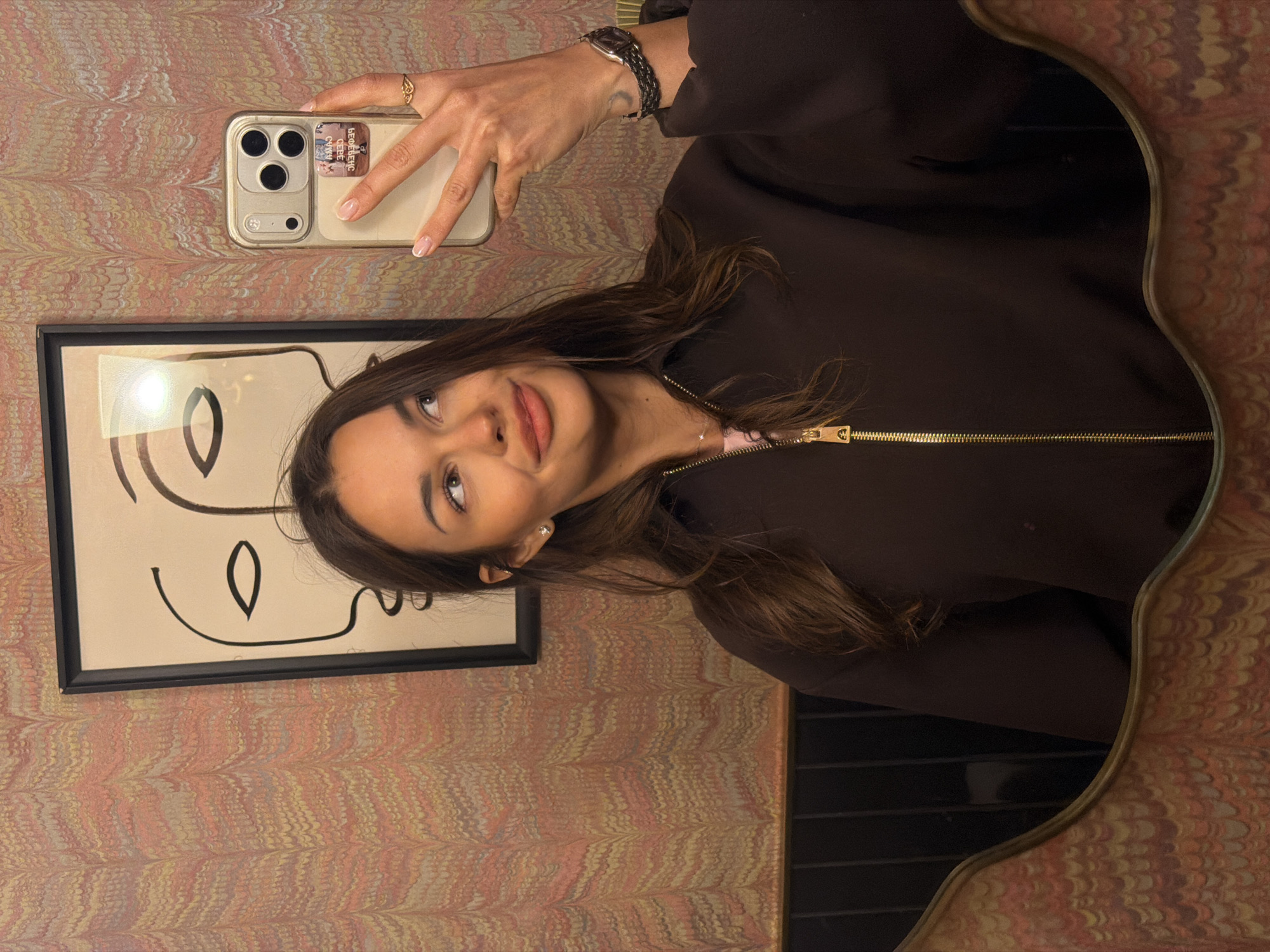 A young woman with long brown hair is taking a mirror selfie in what appears to be a restroom. She is wearing a dark brown zip-up jacket over a pink top, and a delicate necklace. On her right hand, she wears a ring and a watch. In her left hand, she holds a gold iPhone. The background features a patterned wallpaper with swirling colors of pink, orange, yellow, and white. Above and behind her, a framed abstract artwork depicts two minimalist faces. A portion of the wall behind her is black with vertical lines, and the bottom of the image shows the ornate edge of the mirror and a sliver of what looks like a vanity or counter. There is no indication that this scene is located in Saint Petersburg, Russia.